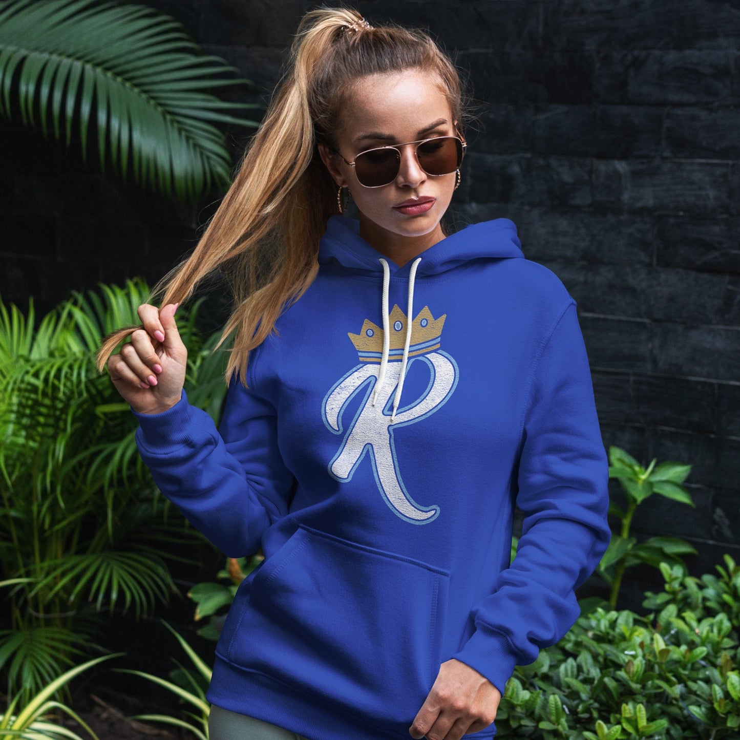 KC Swag Kansas City Royals lite blue/white R wearing gold CROWN on royal blue pull-over hoodie worn by female model standing in front of a dark brick wall and lush green plants