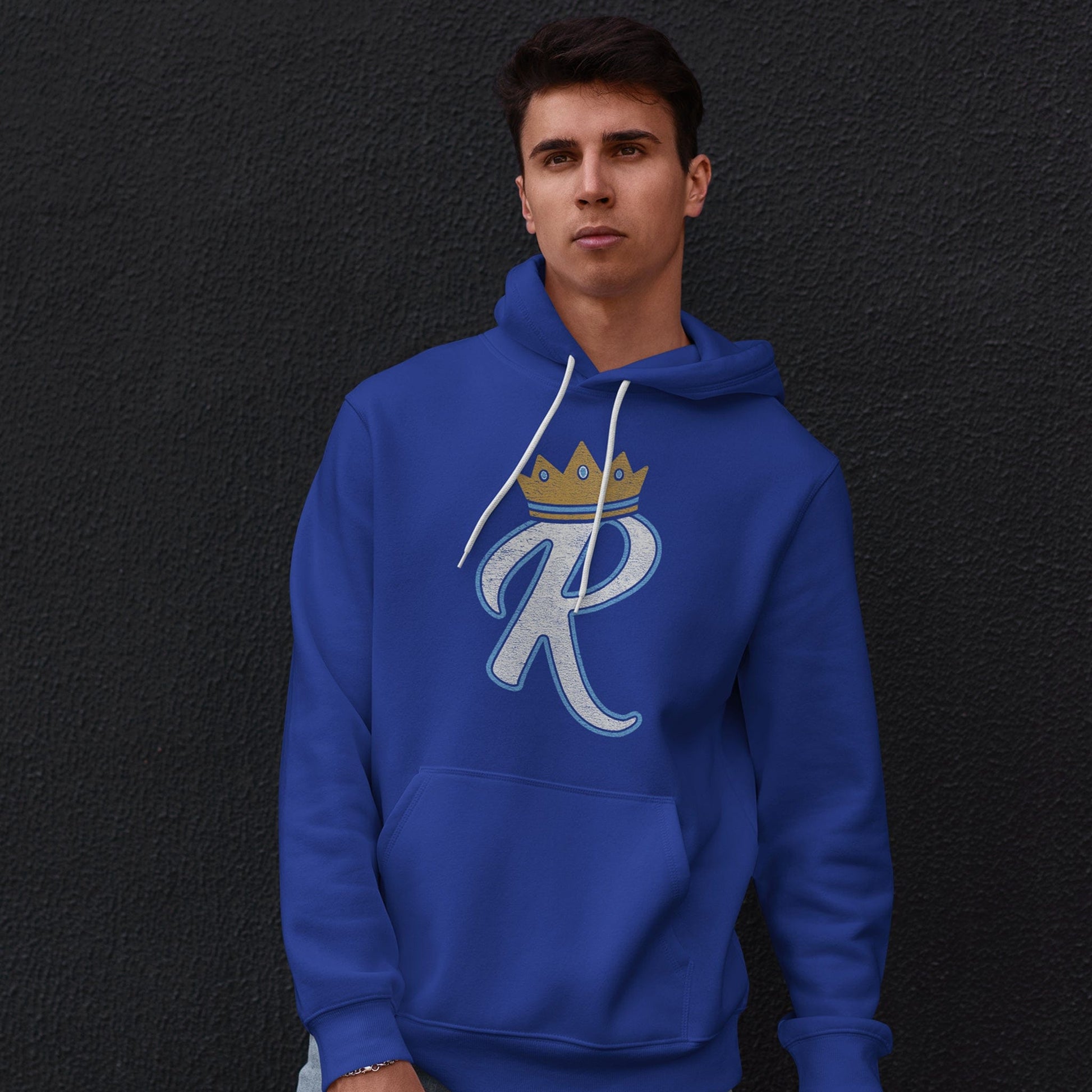 KC Swag Kansas City Royals lite blue/white R wearing gold CROWN on royal blue pull-over hoodie worn by a male model standing in front of a dark grey concrete wall
