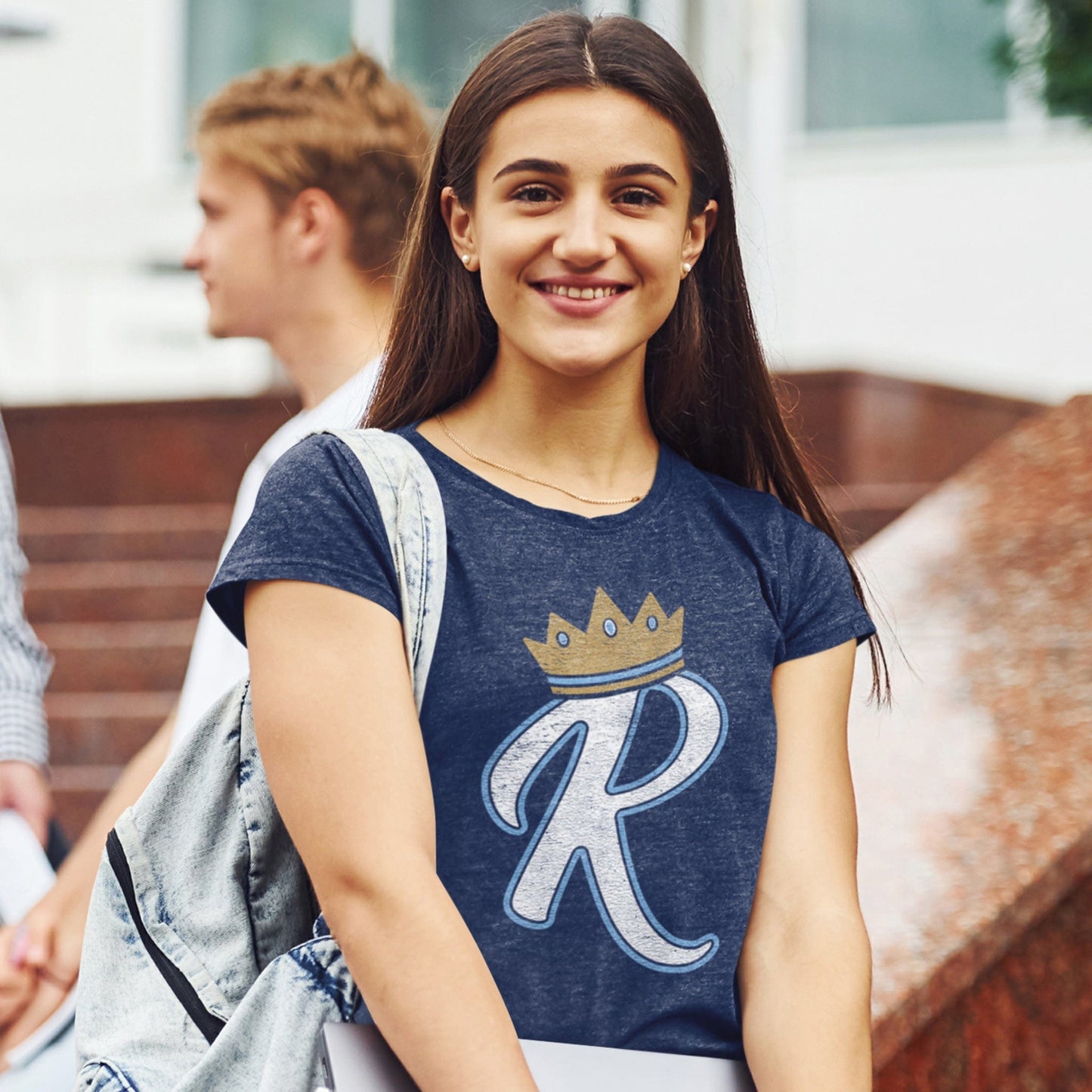 KC Swag Kansas City Royals lite blue/white BOLD R wearing Gold CROWN on heather royal blue t-shirt worn by female model wearing backpack and carrying laptop