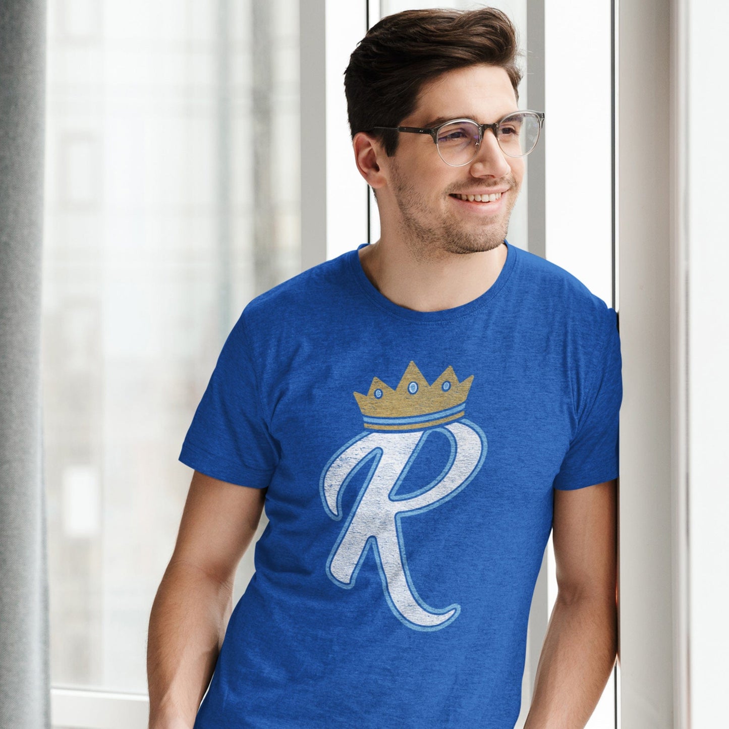KC Swag Kansas City Royals lite blue/white BOLD R wearing Gold CROWN on heather royal blue t-shirt worn by male model leaning against wall near window