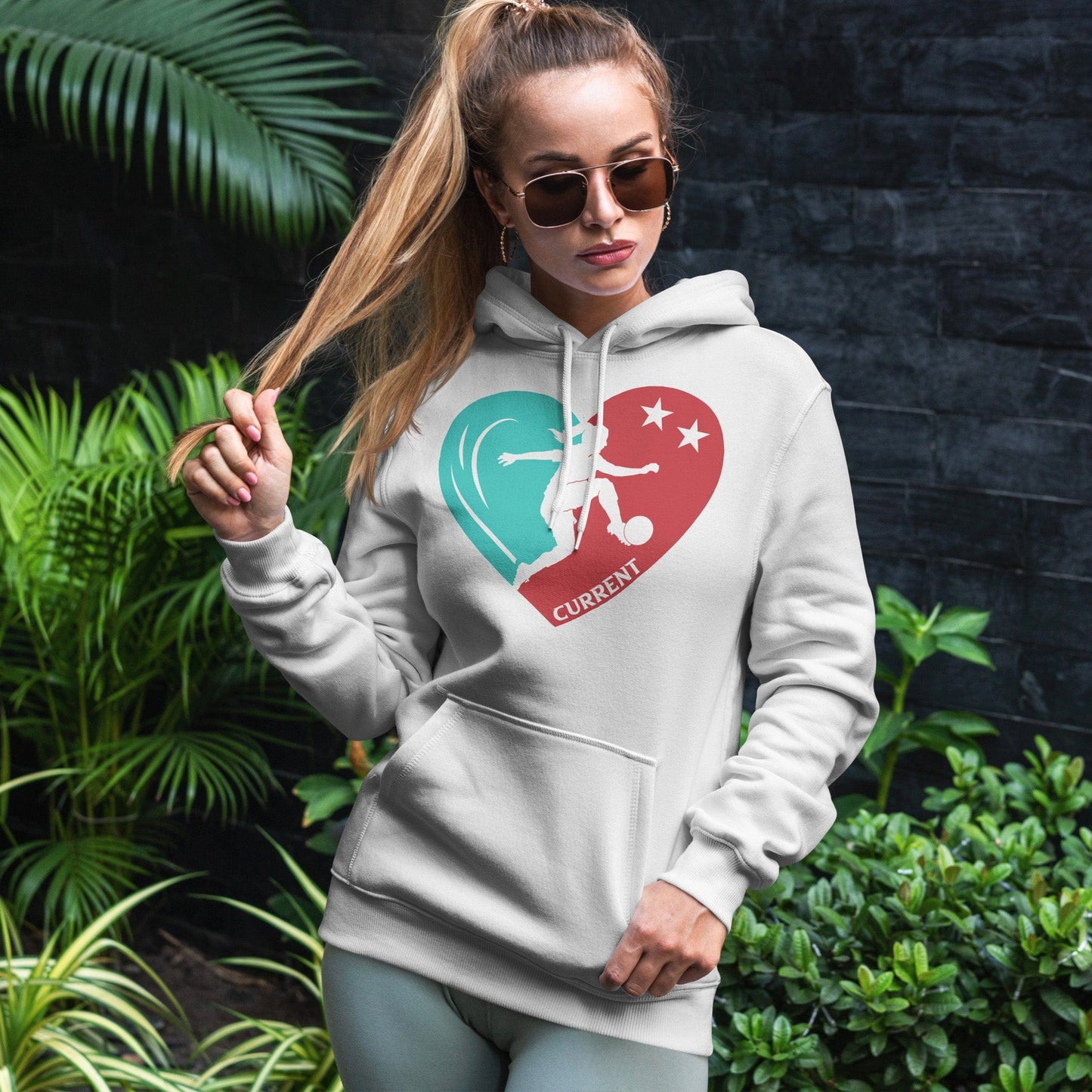 KC Swag Kansas City Current PLAYER HEART on white fleece pullover hoodie worn by female model standing in front of a dark grey brick wall and green foliage.