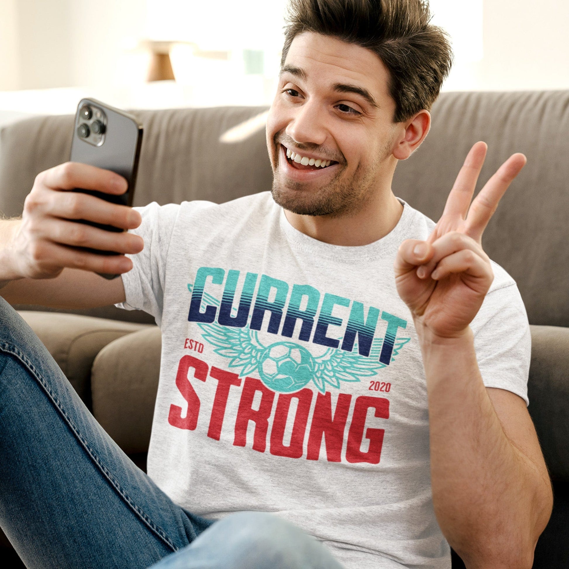 KC Swag Kansas City Current CURRENT STRONG on heather Ash unisex t-shirt worn by male model seated in front of a couch flashing a smile and the peace sign to his mobile phone