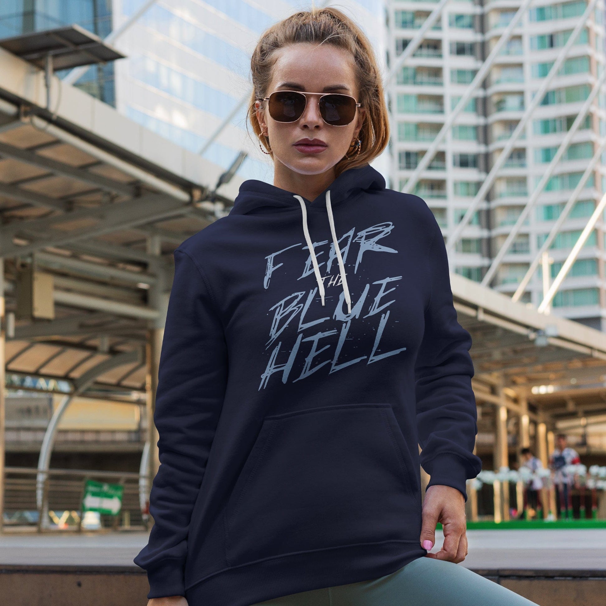KC Swag Sporting Kansas City powder blue/white FEAR THE BLUE HELL on navy fleece pull-over hoodie worn by female model in front of downtown transportation facility