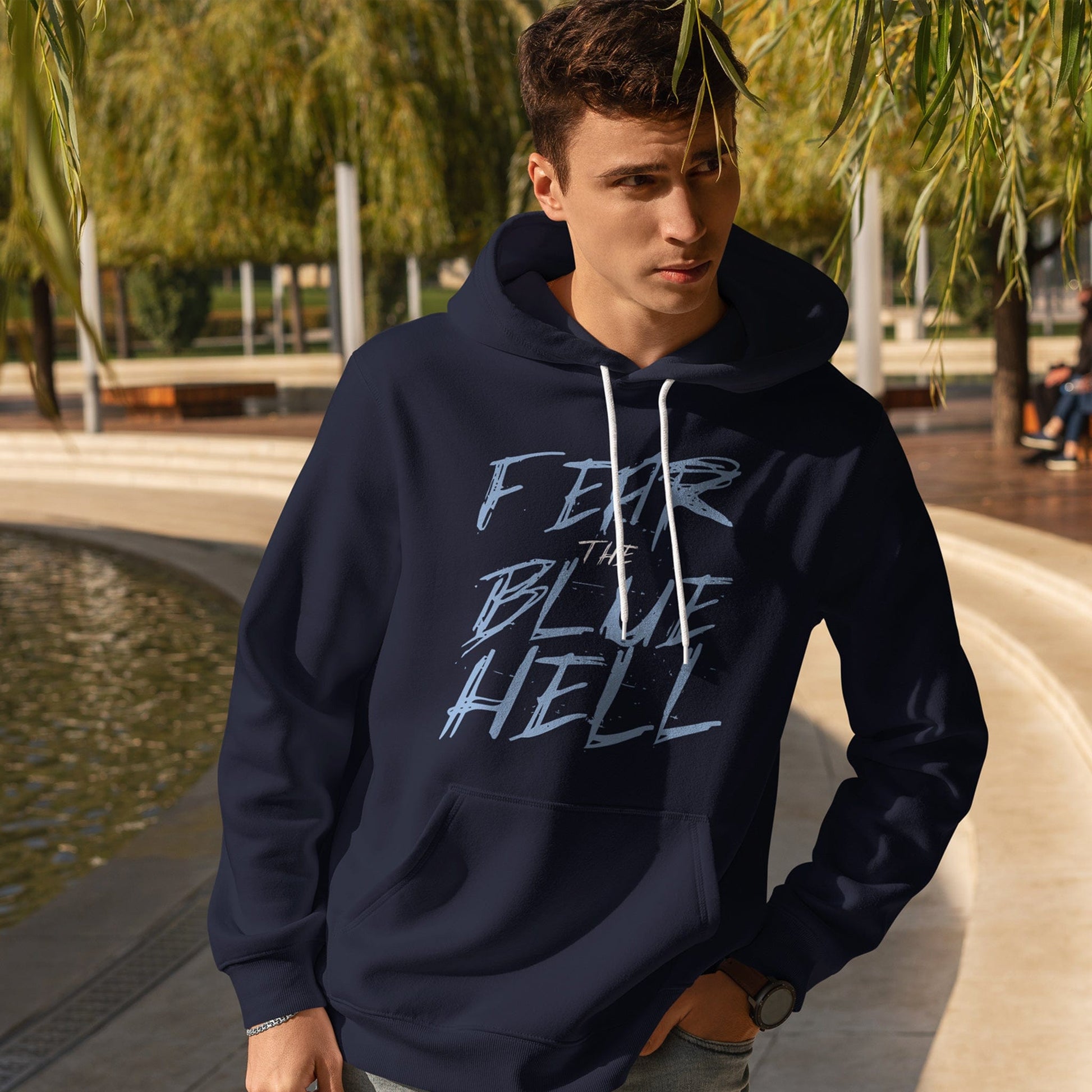 KC Swag Sporting Kansas City powder blue/white FEAR THE BLUE HELL on navy fleece pull-over hoodie worn by male model near water in a city park