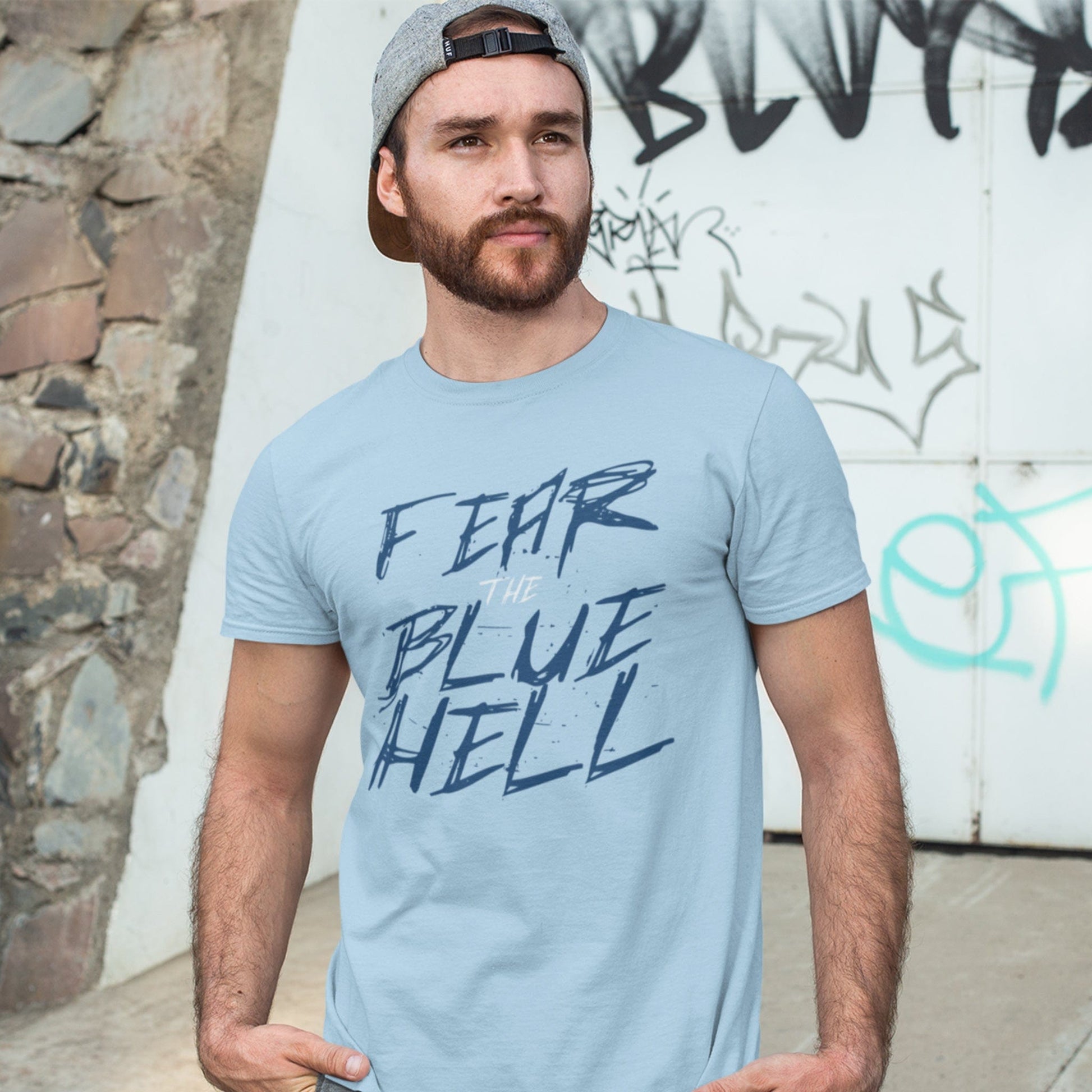 KC Swag Sporting Kansas City navy blue white FEAR THE BLUE HELL on light blue t-shirt worn by male model wearing hat in front of graffiti wall