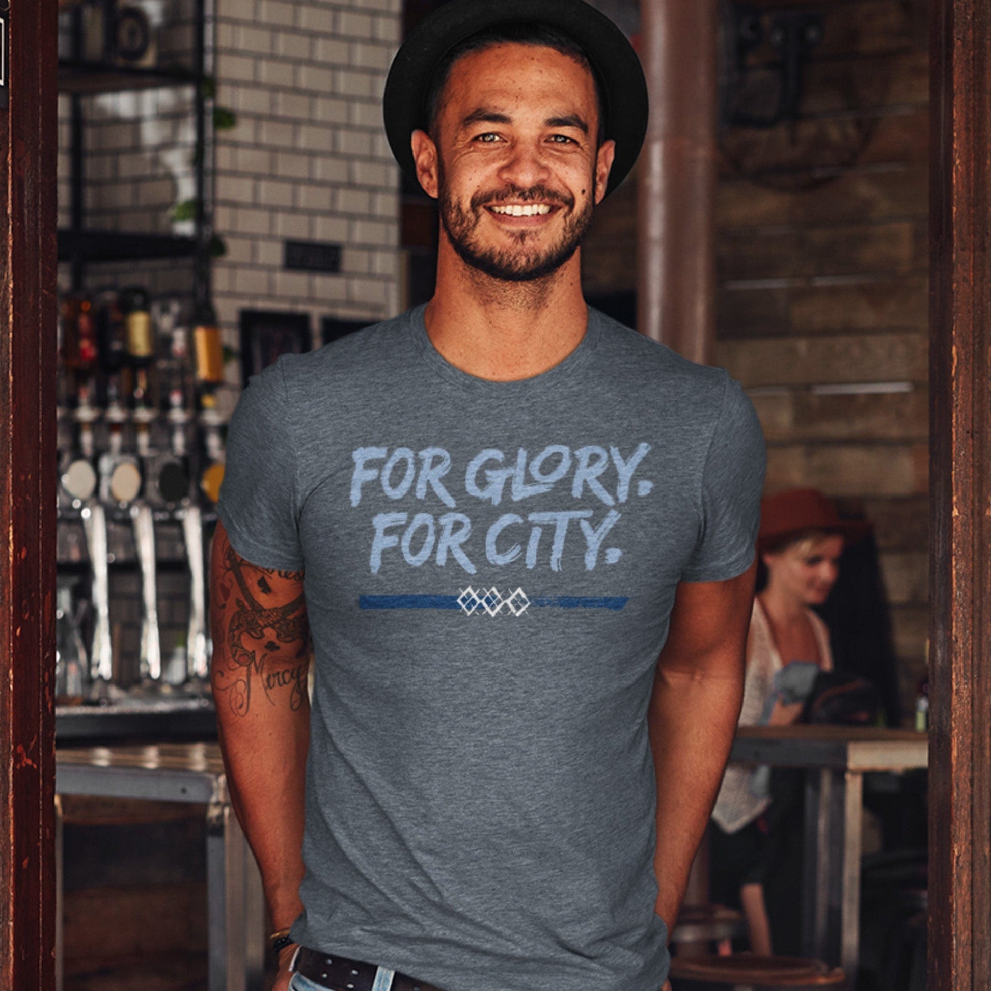 KC Swag Sporting Kansas City navy, powder, white FOR GLORY FOR CITY on heather slate unisex t-shirt worn by male model  standing in doorway to pub
