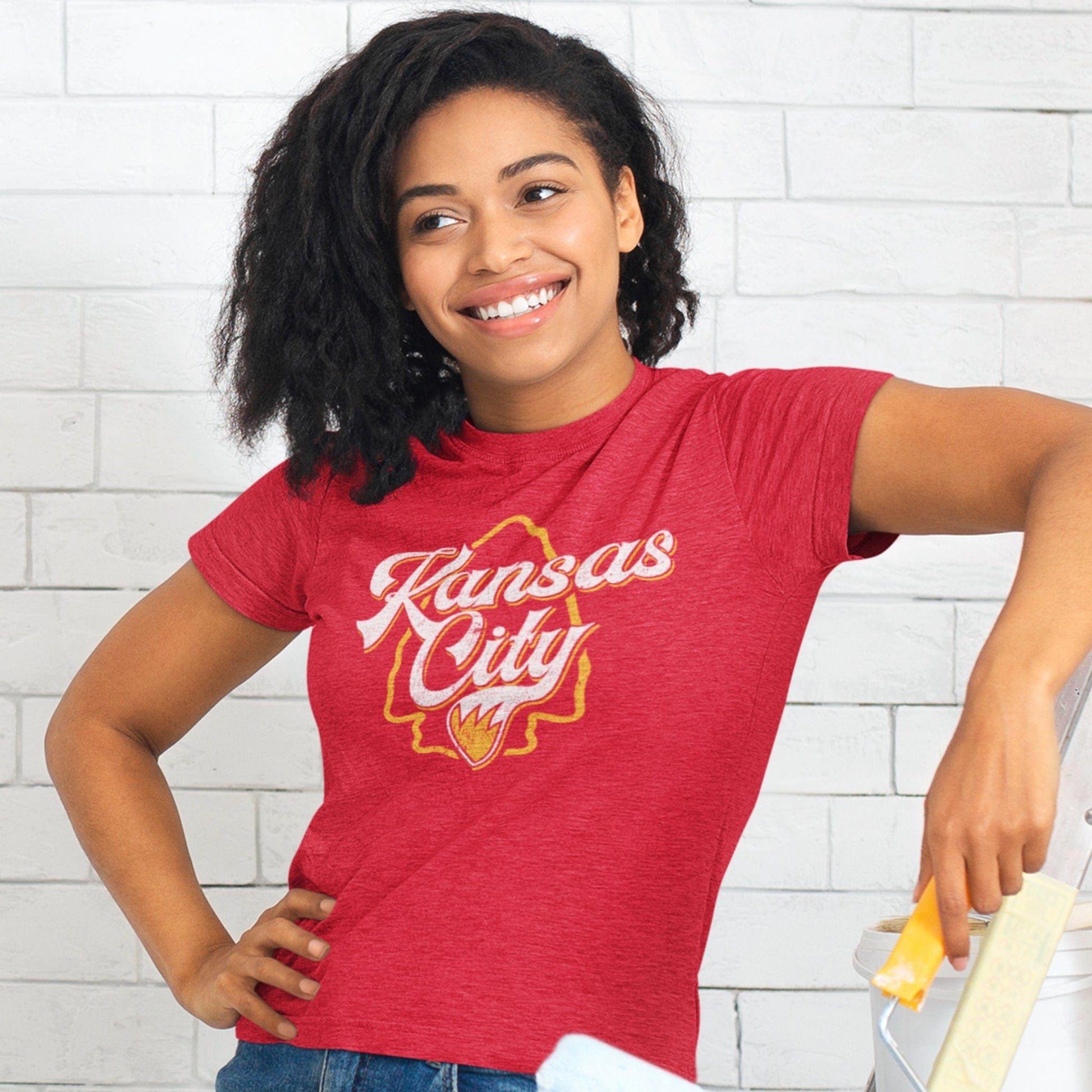 KC Swag Kansas City Chiefs red/yellow KANSAS CITY (with foxtail) with arrowhead graphic on heather red t-shirt worn by female model painting a white brick wall
