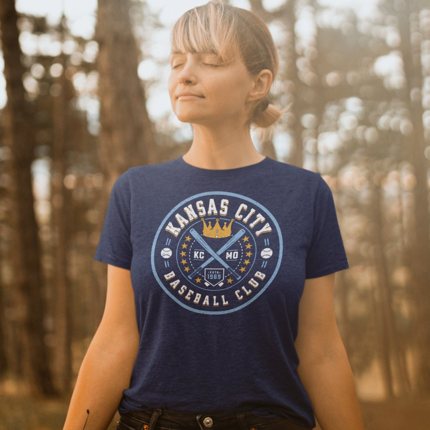 KC Swag Kansas City Royals blue, gold, white KC BASEBALL CLUB on heather navy unisex t-shirt worn by female model standing in warm wooded area
