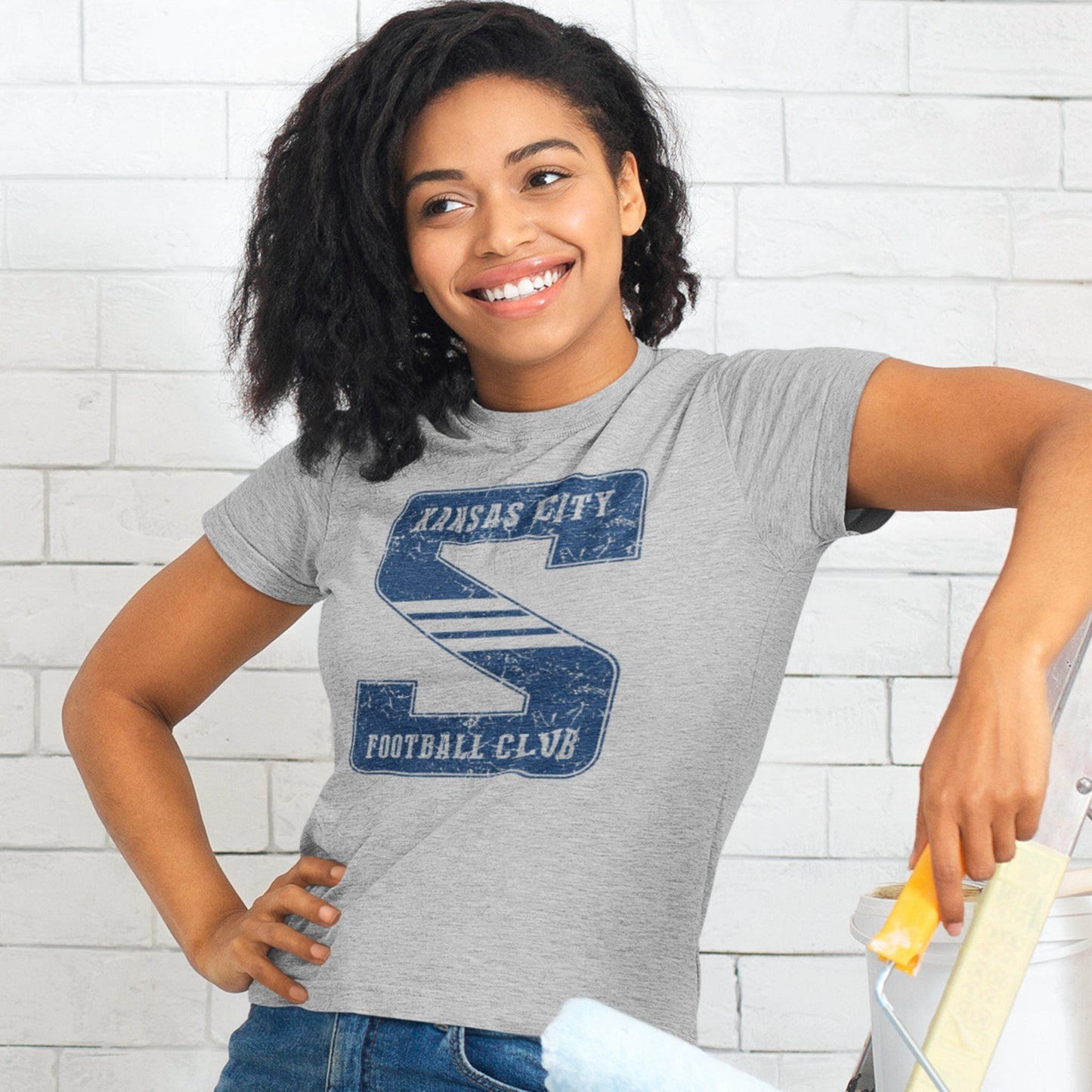 KC Swag Sporting Kansas City navy KANSAS CITY FOOTBALL CLUB on giant striped S on athletic heather grey t-shirt worn by female model painting a white brick wall