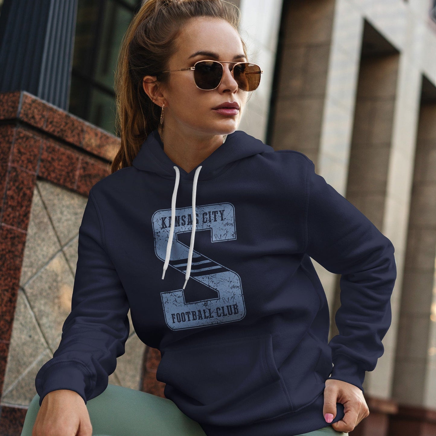 KC Swag Sporting Kansas City powder blue KANSAS CITY FOOTBALL CLUB in STRIPED S on navy fleece pull-over hoodie worn by female model sitting in front of stone building