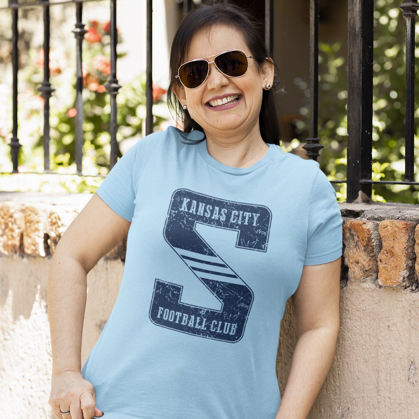 KC Swag Sporting Kansas City navy KANSAS CITY FOOTBALL CLUB on giant striped S on lite blue t-shirt worn by female model leaning on concrete wall wearing sunglasses