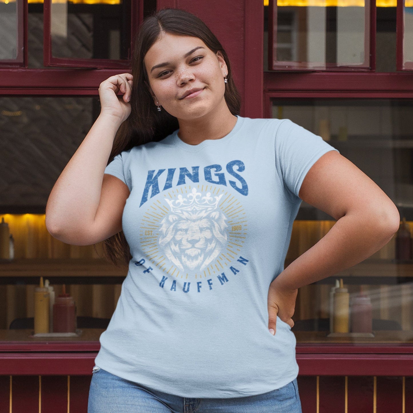 KC Swag Kansas City Royals blue/white/gold KINGS OF KAUFFMAN with lion wearing crown on light blue t-shirt worn by female model standing in front of restaurant windows