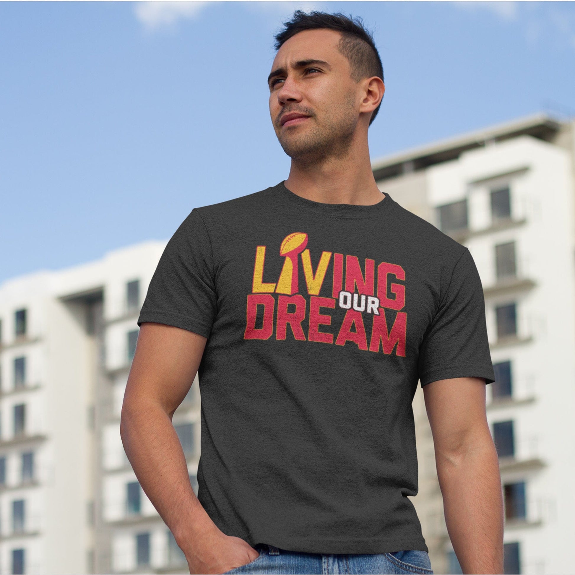 KC Swag Kansas City Chiefs red/yellow/white L(Lombardi trophy)IVING OUR DREAM on dark heather grey t-shirt worn by male model in front of apartment building