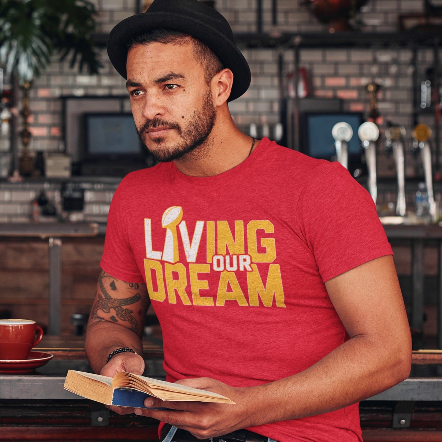 KC Swag Kansas City Chiefs red/yellow/white L(Lombardi trophy)IVING OUR DREAM on heather red t-shirt worn by male model sitting in cafe