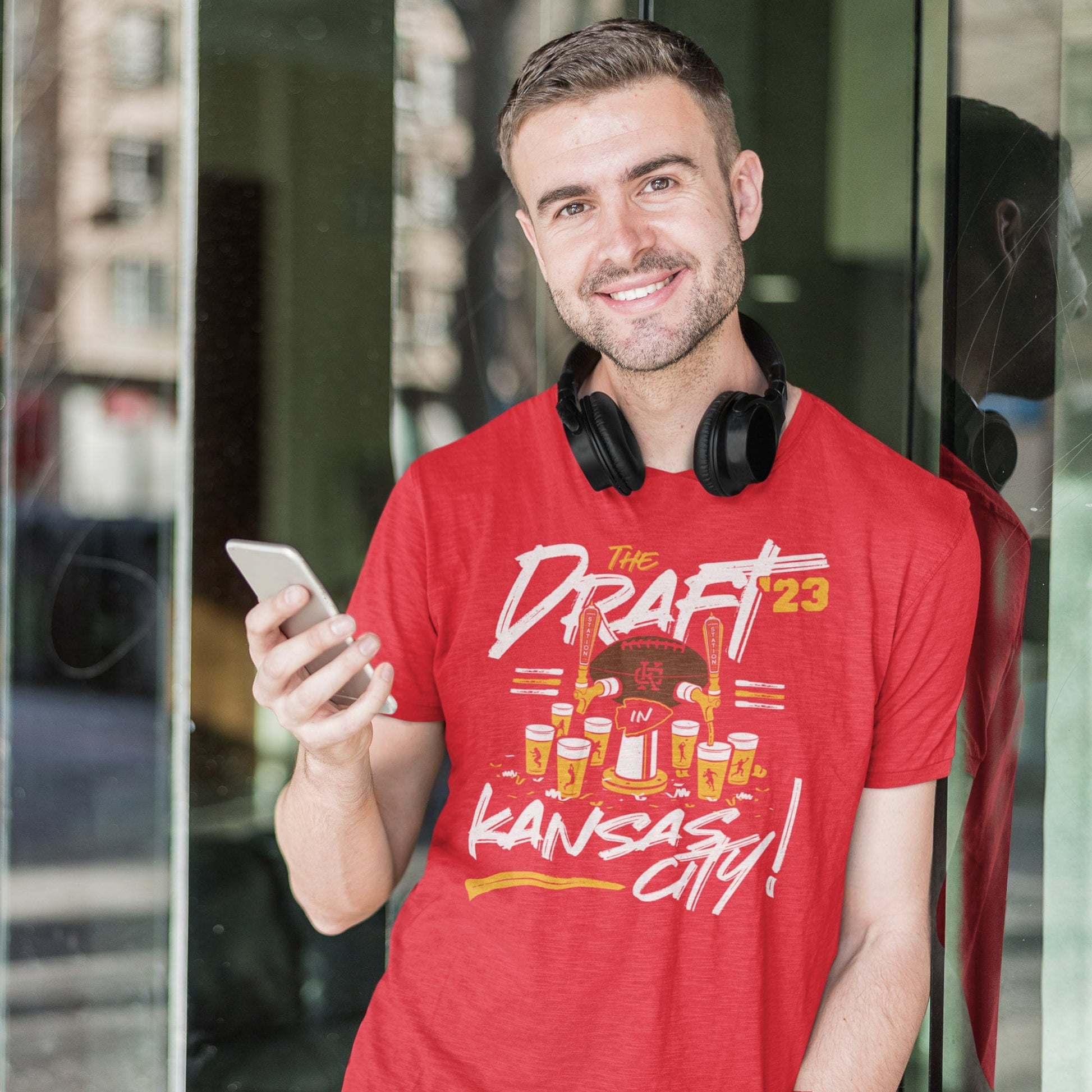 KC Swag Kanssas City Chiefs DRAFTIN' IN KC on heather red unisex t-shirt worn by male model leaning against large glass window holding his phone