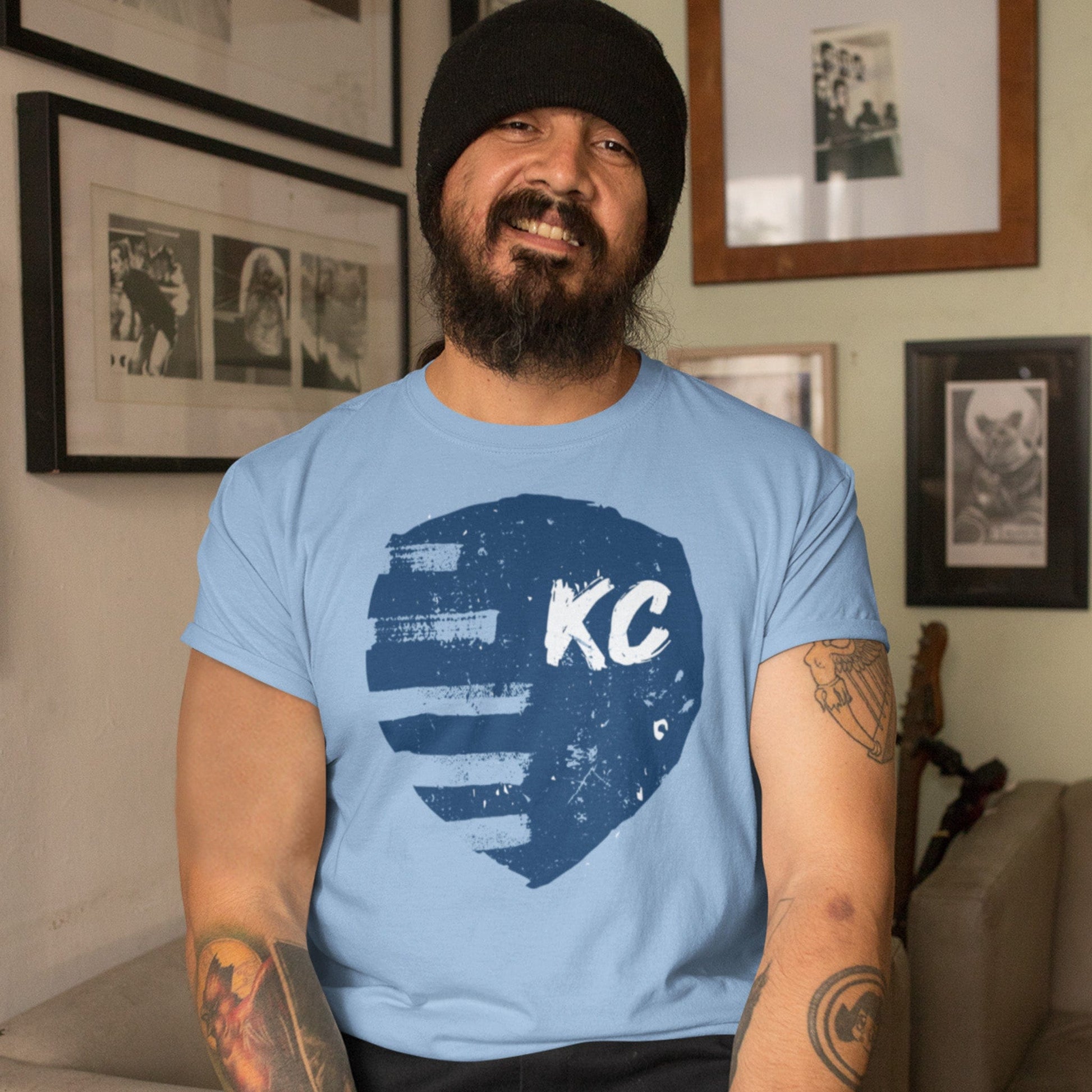 KC Swag Sporting Kansas City navy, powder, white PAINTED SHIELD on baby blue unisex t-shirt worn by male model sitting in living room in front of wall of pictures