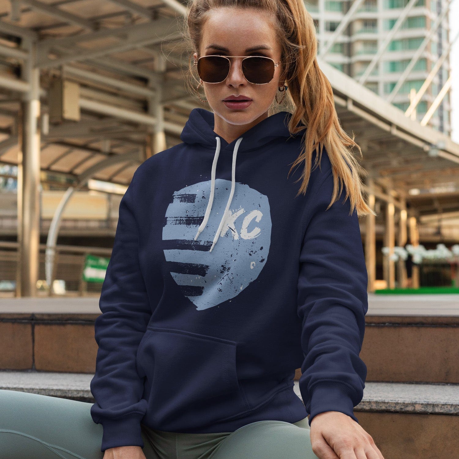 KC Swag Sporting Kansas City powder, white PAINTED SHIELD on navy fleece pullover hoodie worn by female model sitting in front of downtown bus terminal