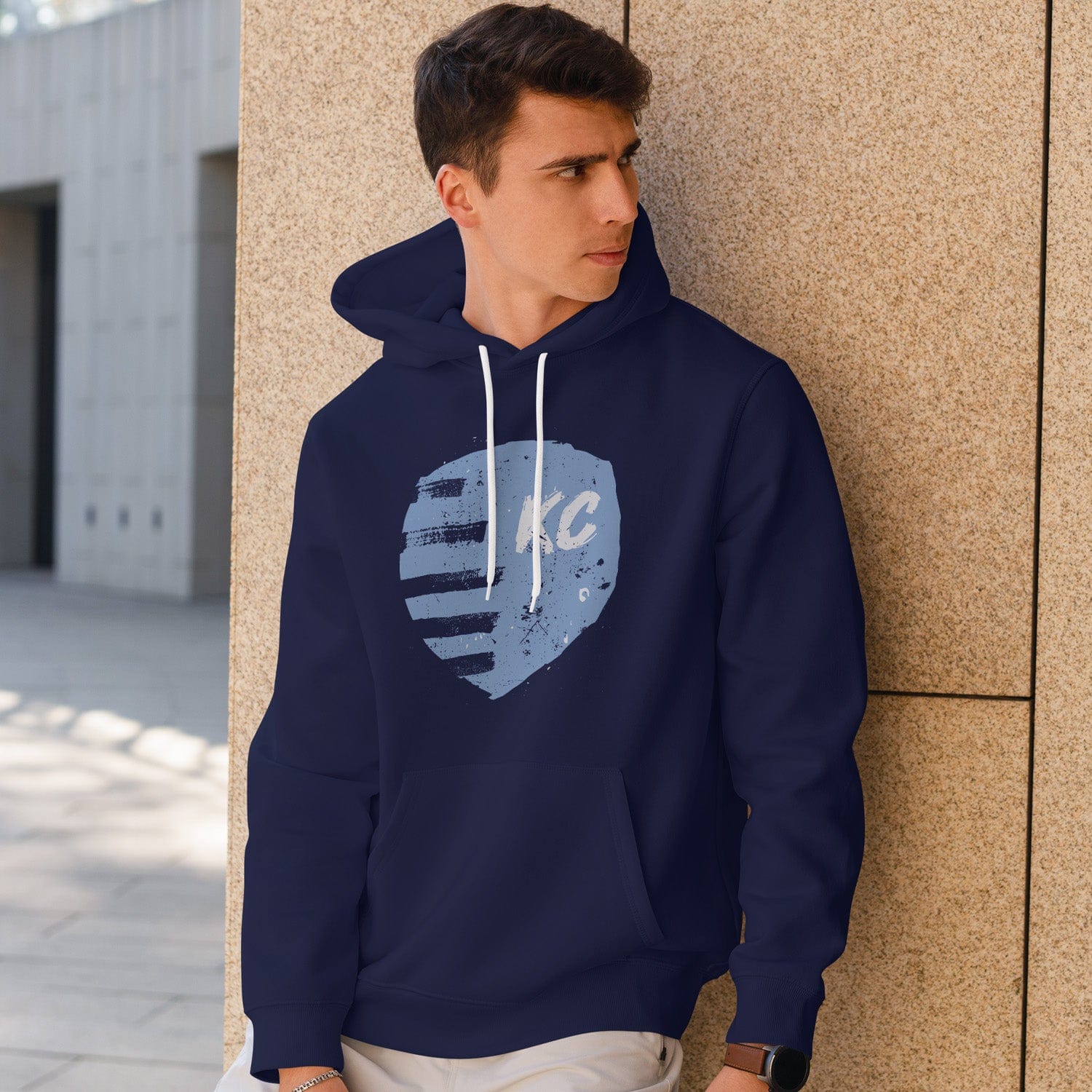 KC Swag Sporting Kansas City powder, white PAINTED SHIELD on navy fleece pullover hoodie worn by male model leaning on stone wall in outdoor plaza