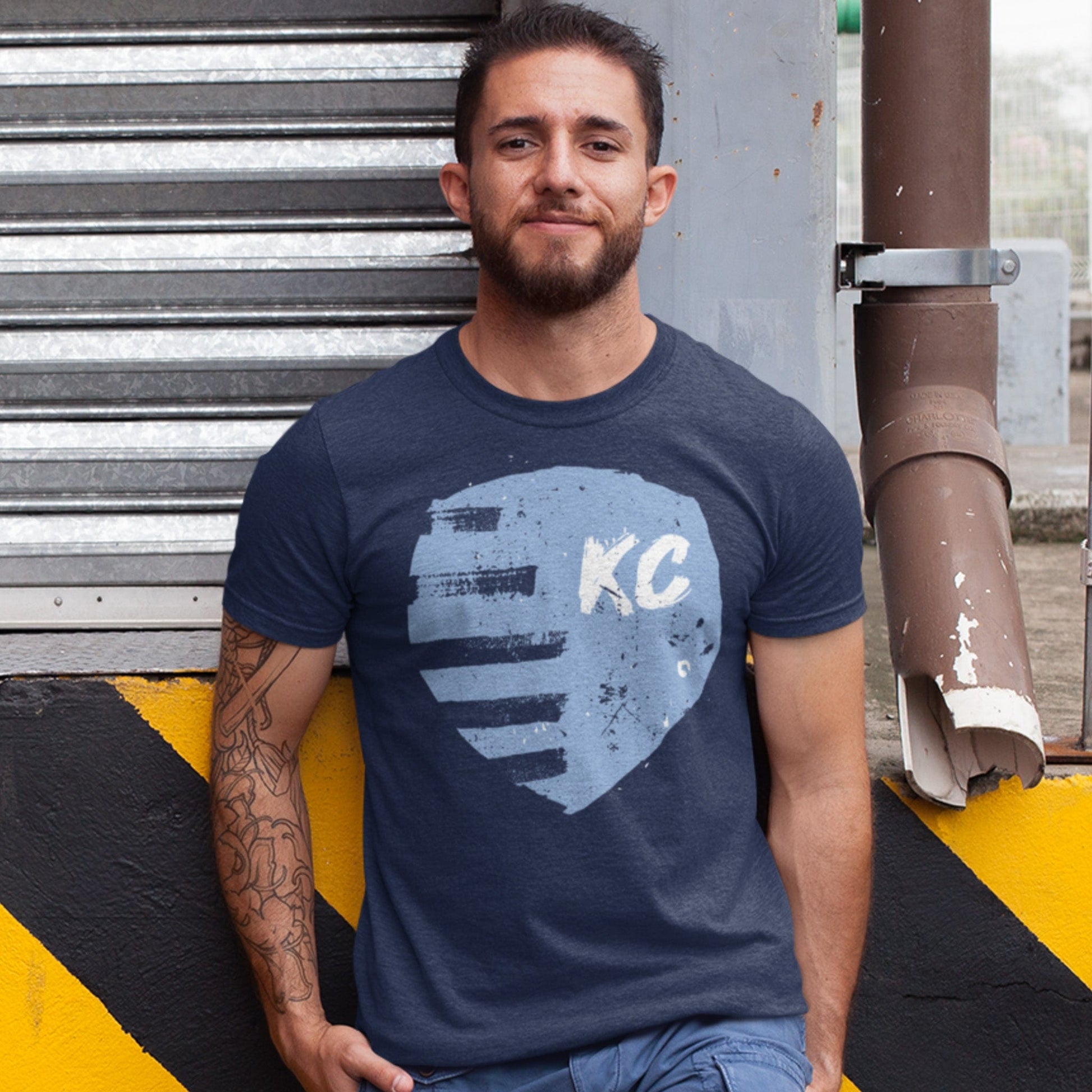 KC Swag Sporting Kansas City navy, powder, white PAINTED SHIELD on heather navy unisex t-shirt worn by male model leaning on loading dock in urban setting