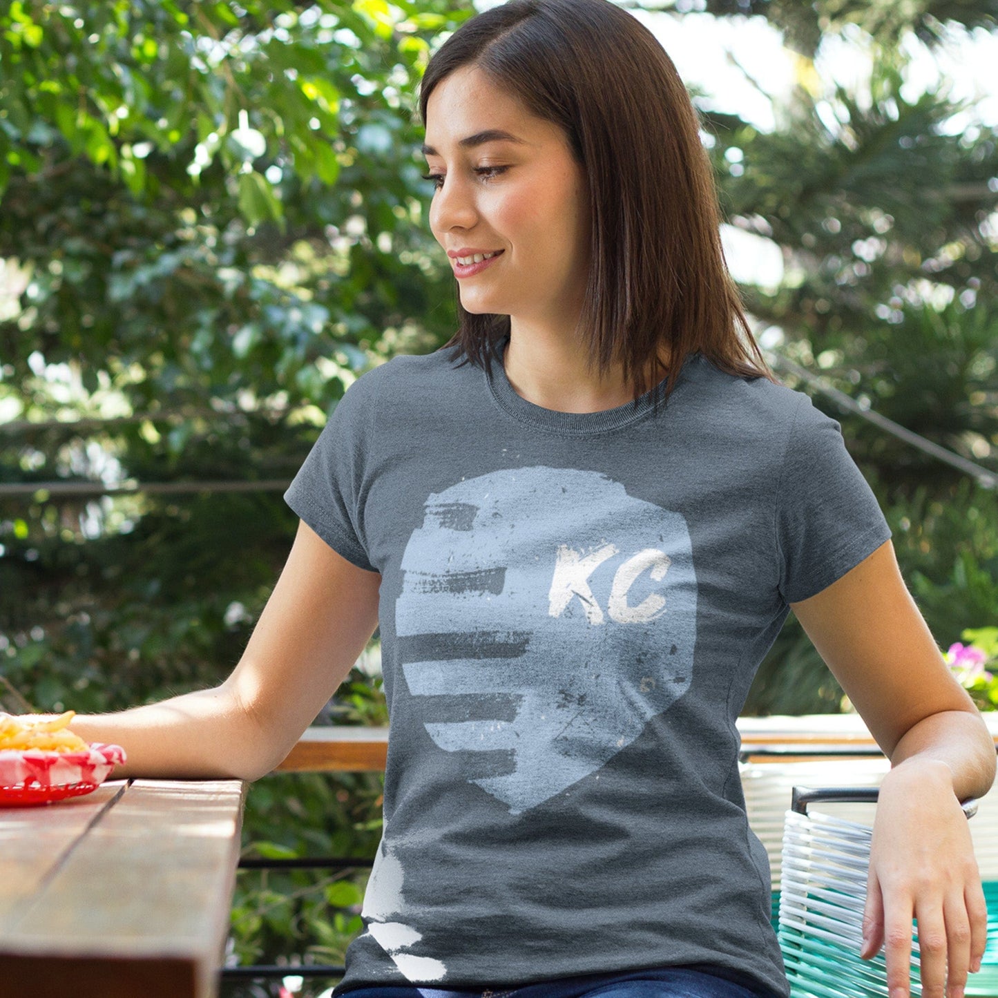 KC Swag Sporting Kansas City navy, powder, white PAINTED SHIELD on heather slate unisex t-shirt worn by female model sitting in picnic table on outdoor patio