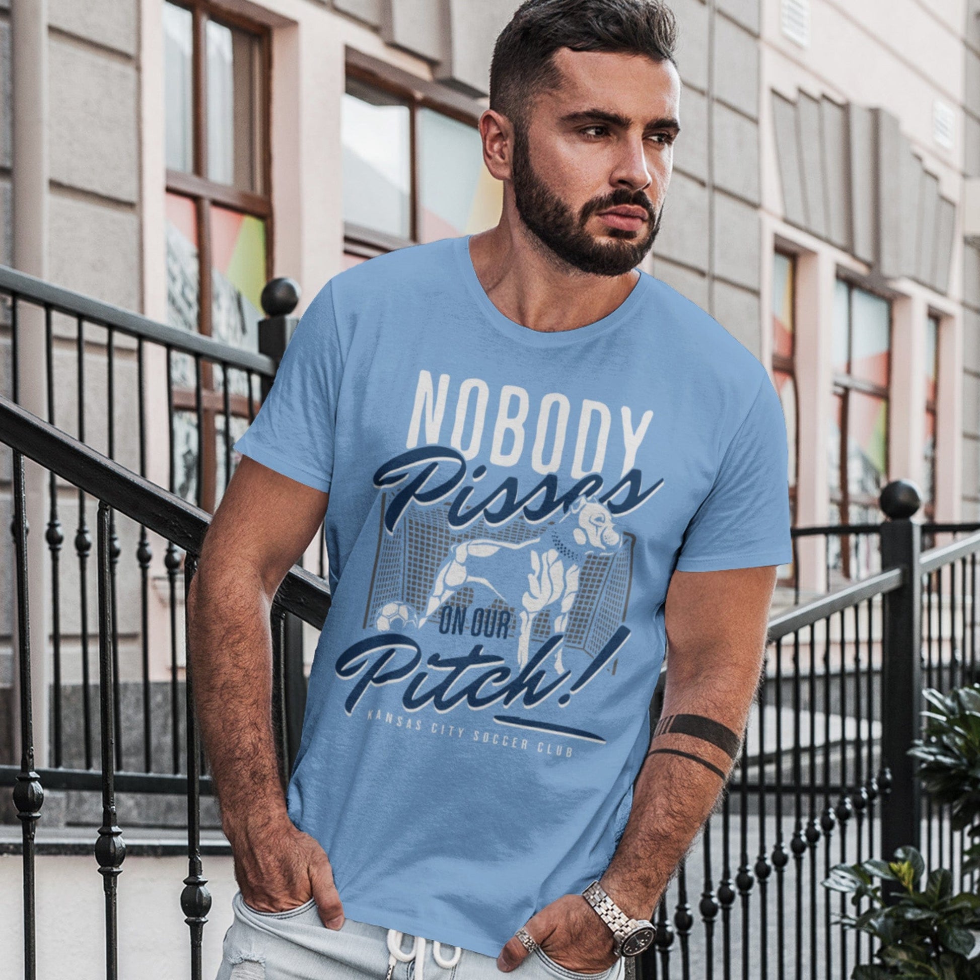 KC Swag Sporting Kansas City navy, powder, white, grey PITCH PROTECTOR on baby blue unisex t-shirt worn by male model standing on apartment stoop