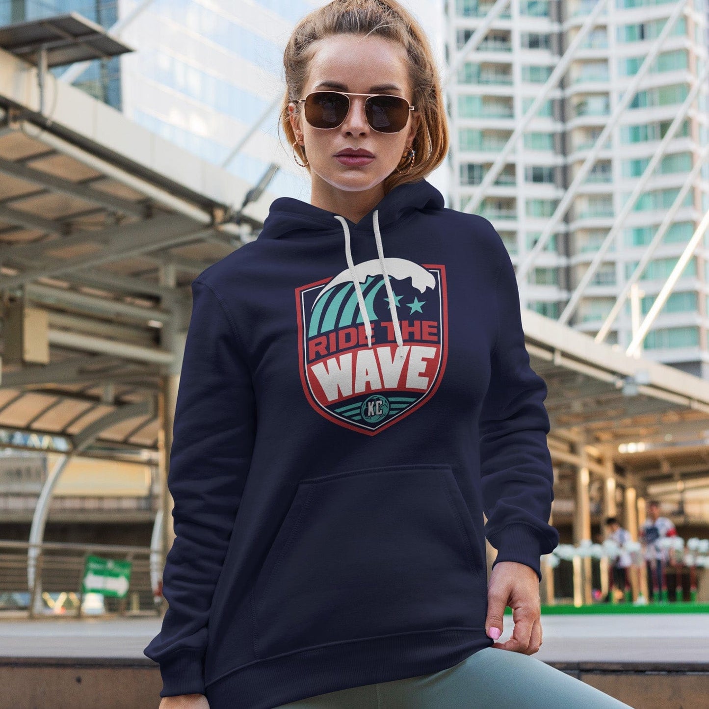 KC Swag Kansas City Current RIDE THE WAVE on navy fleece pullover hoodie worn by female model standing in front of downtown transit station