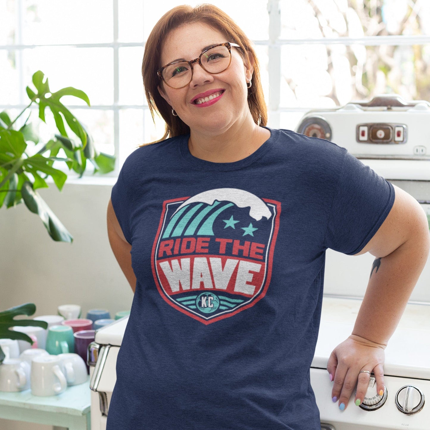 KC Swag Kansas City Current RIDE THE WAVE on heather navy unisex t-shirt worn by female model leaning on stove in bright kitchen