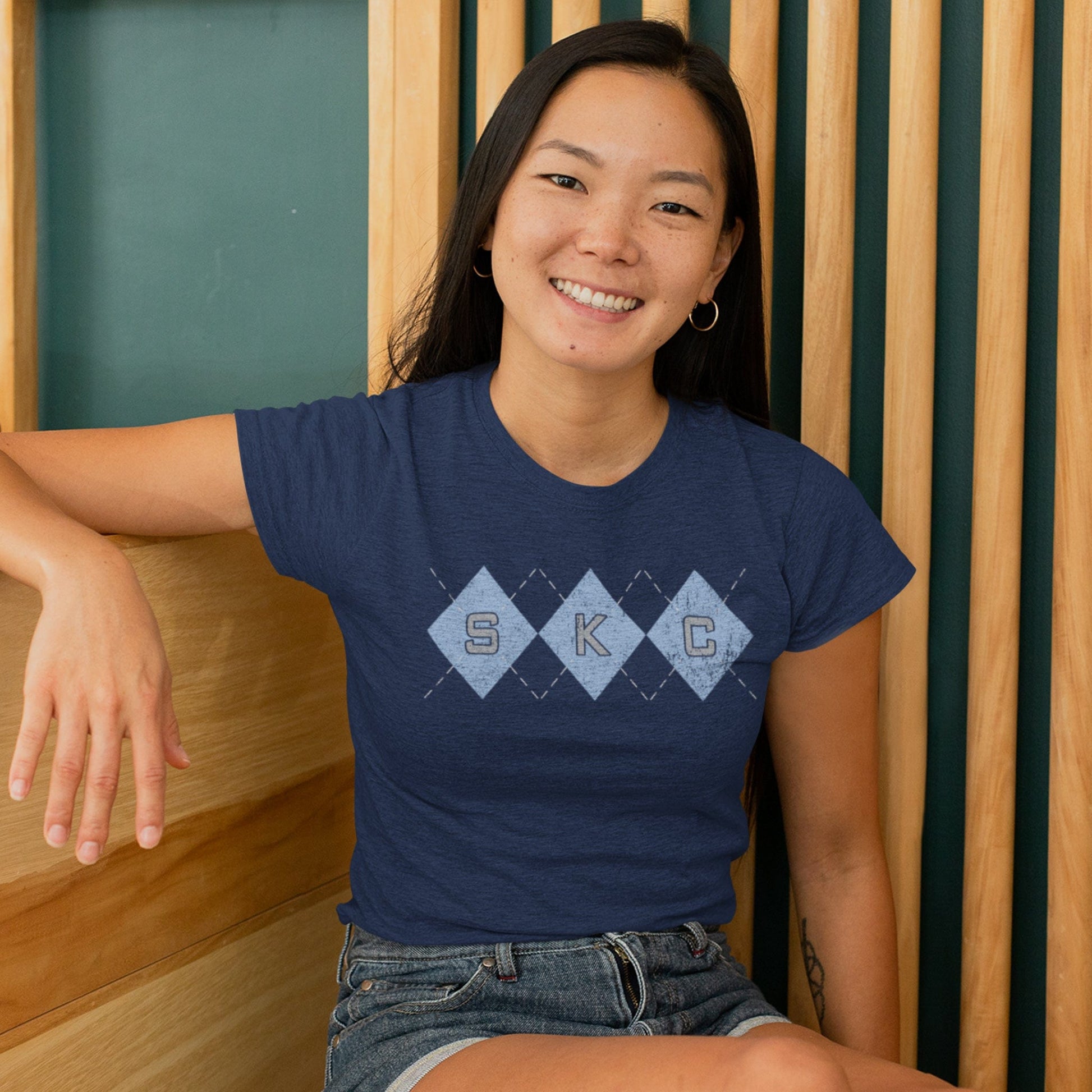 KC Swag Sporting Kansas City powder blue/grey SKC DIAMONDS with argyle stitching on heather navy t-shirt worn by female model sitting on wood bench in front of wood slats