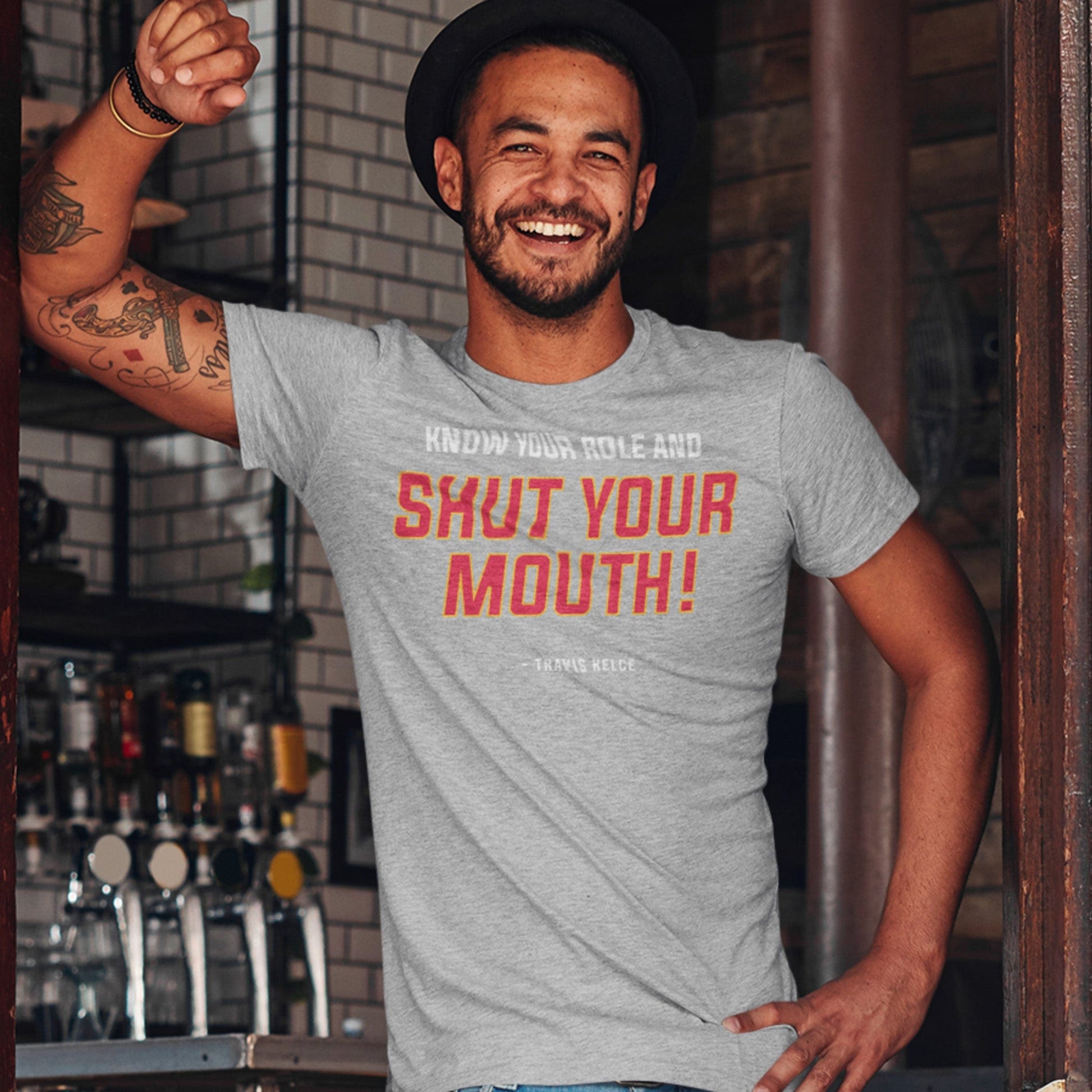 KC Swah Kansas City Chiefs red, yellow, white SHUT YOUR MOUTH on athletic heather grey unisex t-shirt worn by male model standing in door of pub