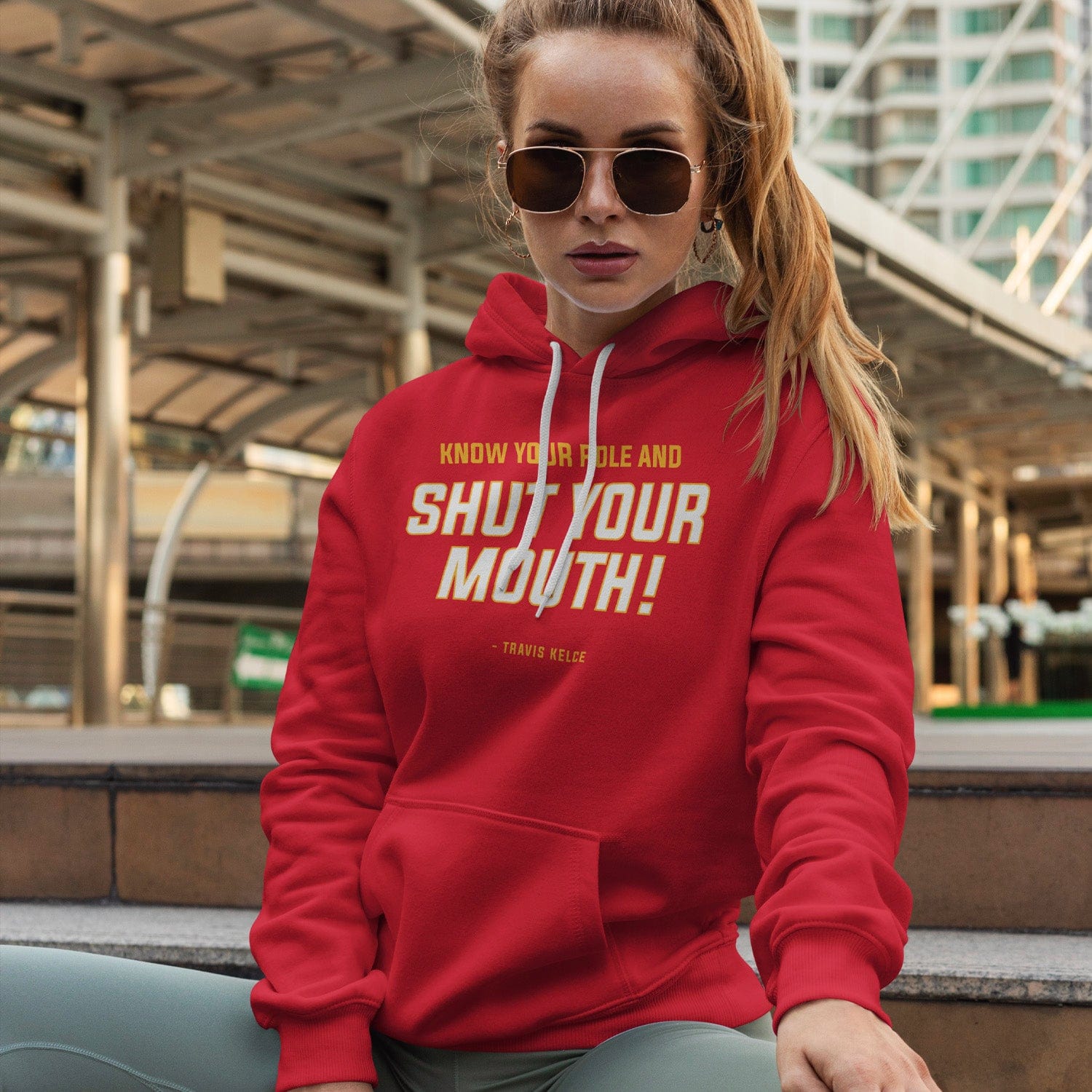 KC Swag Kansas City Chiefs whute, yellow SHUT YOUR MOUTH on red fleece pullover hoodie worn by female model sitting in front of downtown transportation terminal