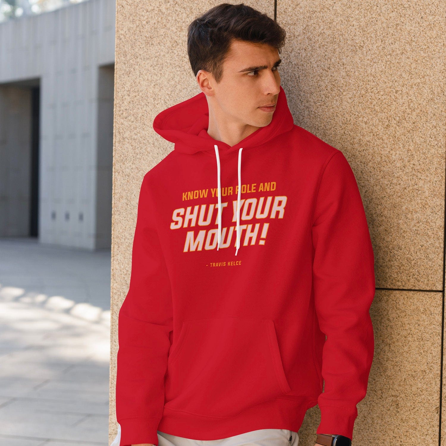 KC Swag Kansas City Chiefs whute, yellow SHUT YOUR MOUTH on red fleece pullover hoodie worn by male model leaning on stone wall in outdoor plaza