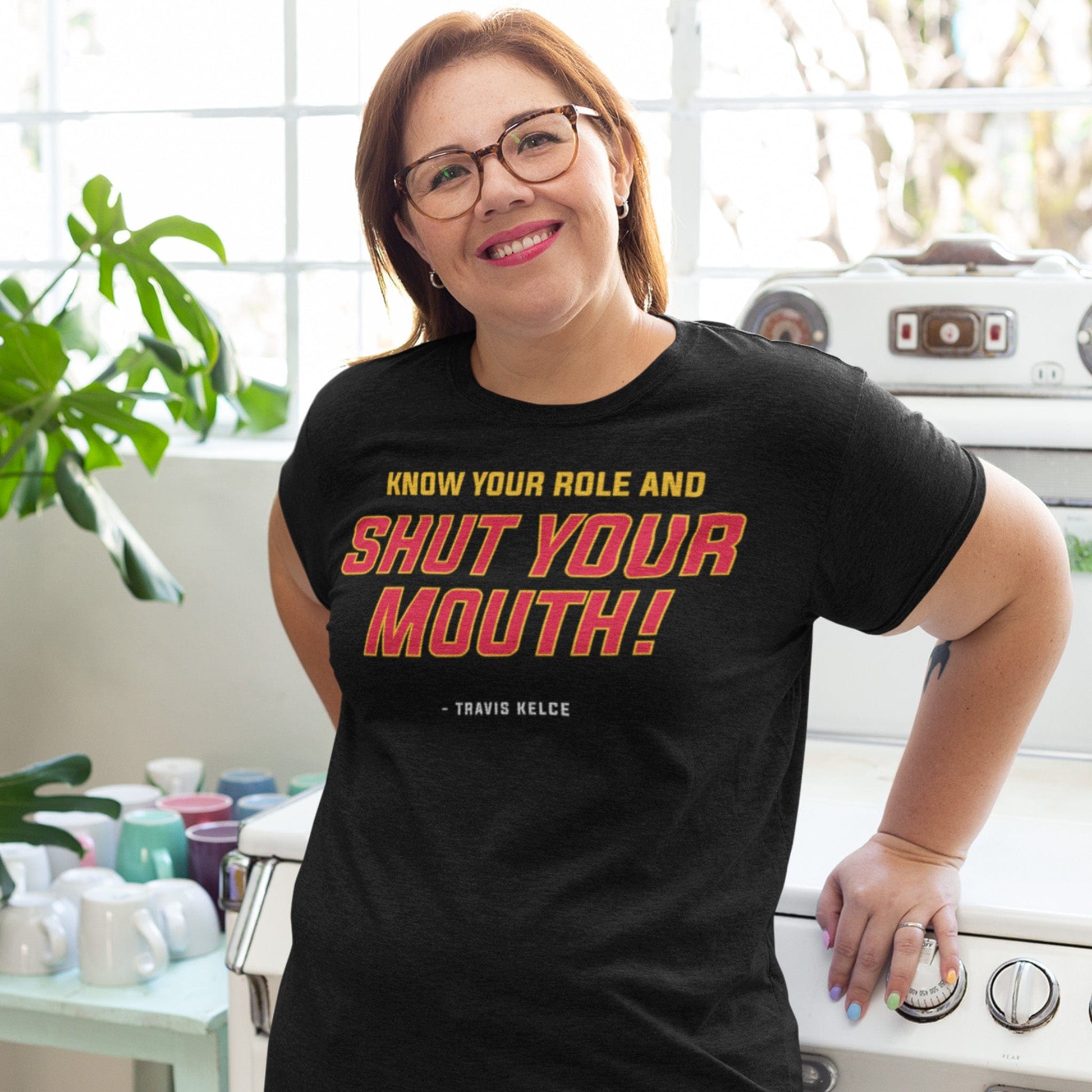 KC Swah Kansas City Chiefs red, yellow, white SHUT YOUR MOUTH on heather black unisex t-shirt worn by female model leaning on stove in bright kitchen