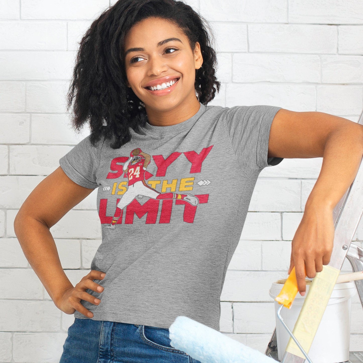 KC Swag Kansas City Chiefs SKYY IS THE LIMIT with football player graphic on athletic heather grey t-shirt worn by female model painting white brick wall