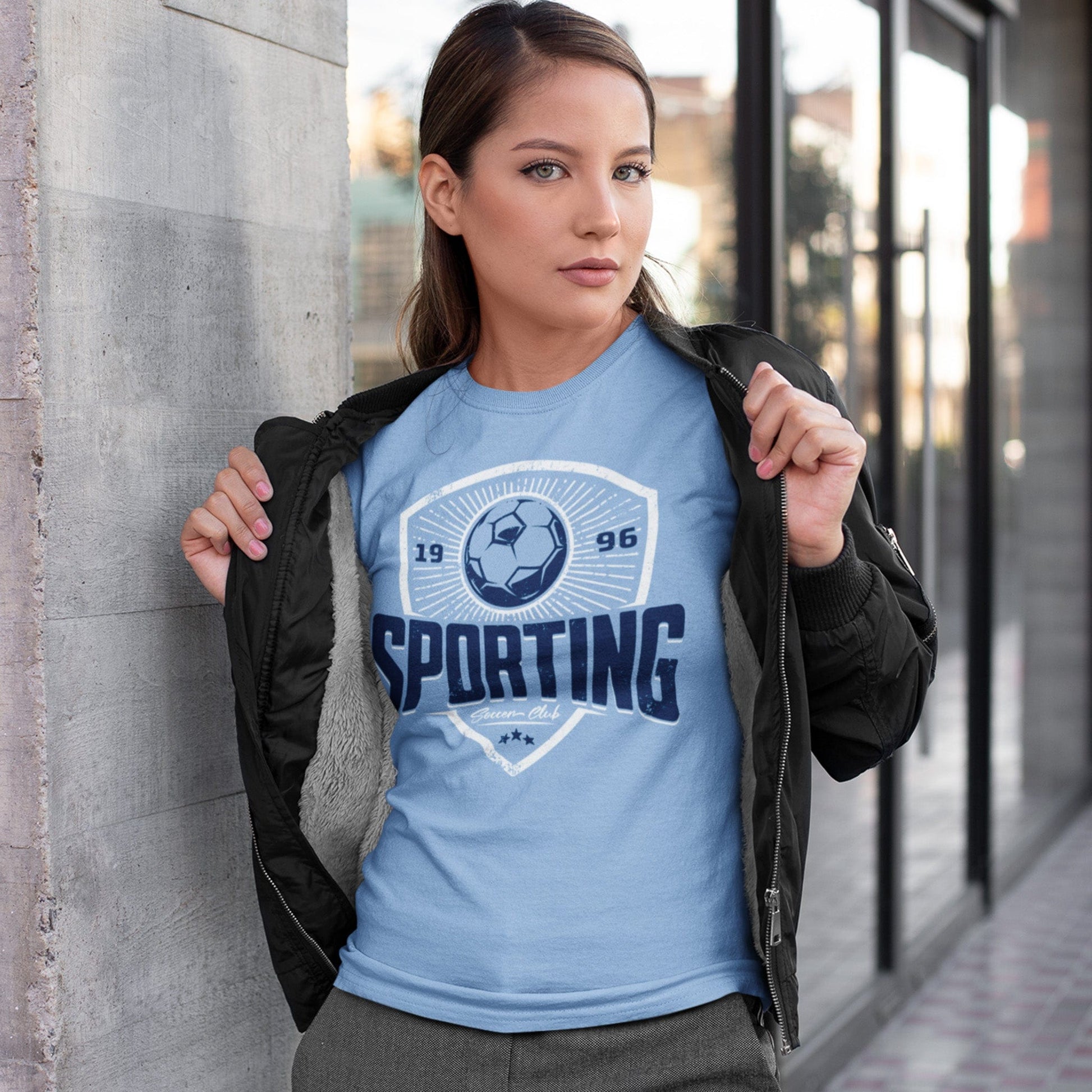 KC Swag Sporting Kansas City navy, powder, white SPORTING CLUB on baby blue unisex t-shirt worn by female model under bomber jacket leaning on concrete wall