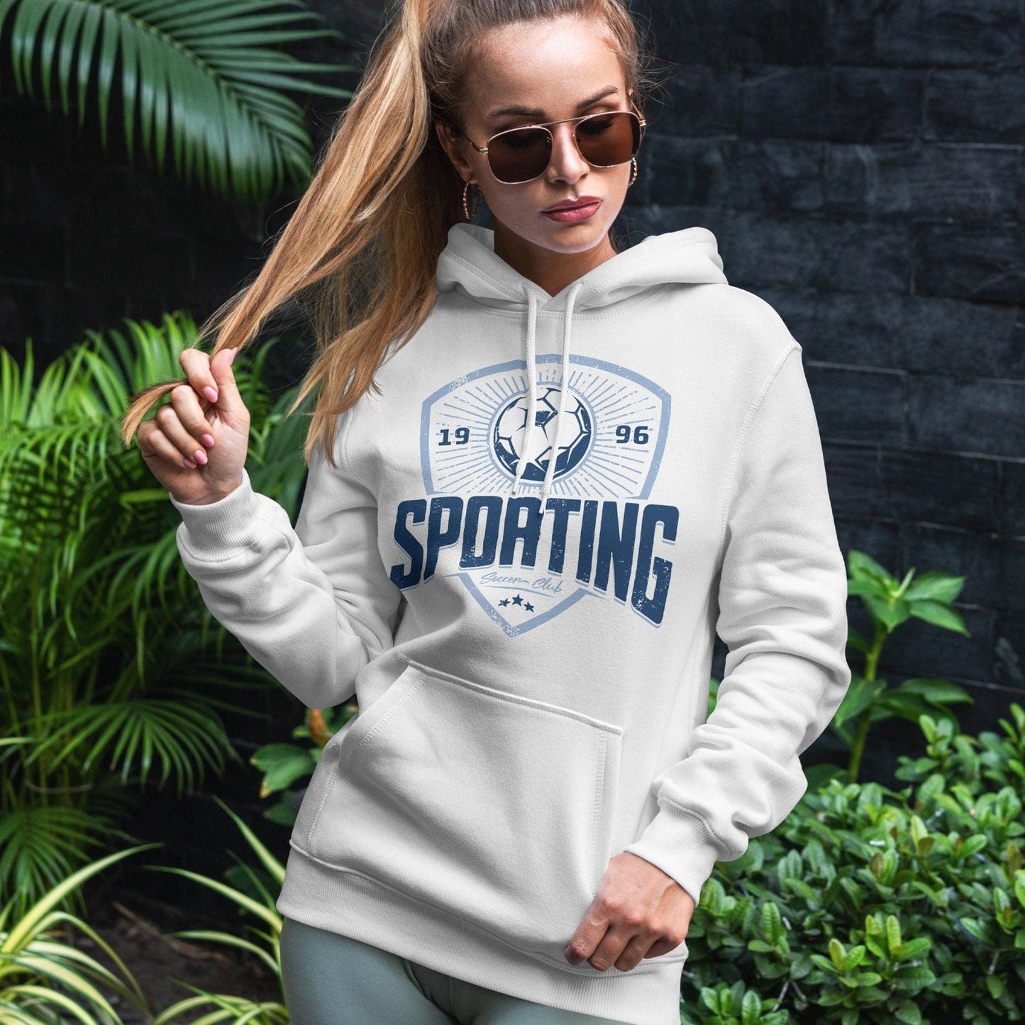 KC Swag Sporting Kansas City navy, powder SPORTING CLUB on white fleece pullover hoodie worn by female model standing in front of dark grey wall and green plants