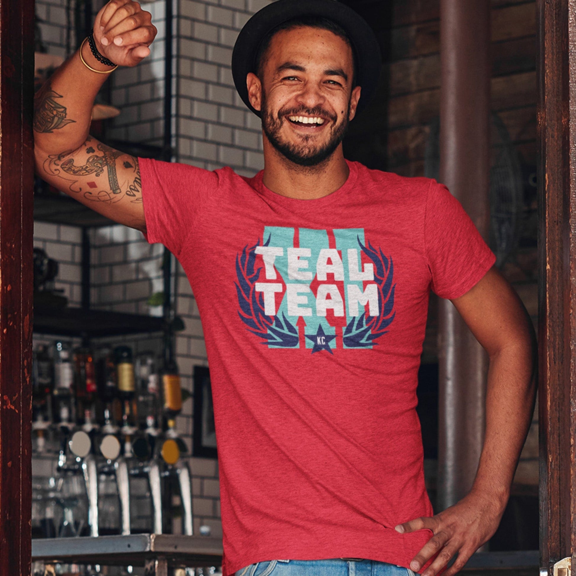 KC Swag Kansas City Current TEAL TEAM X! on heather red unisex t-shirt worn by male model leaning on doorway into pub.