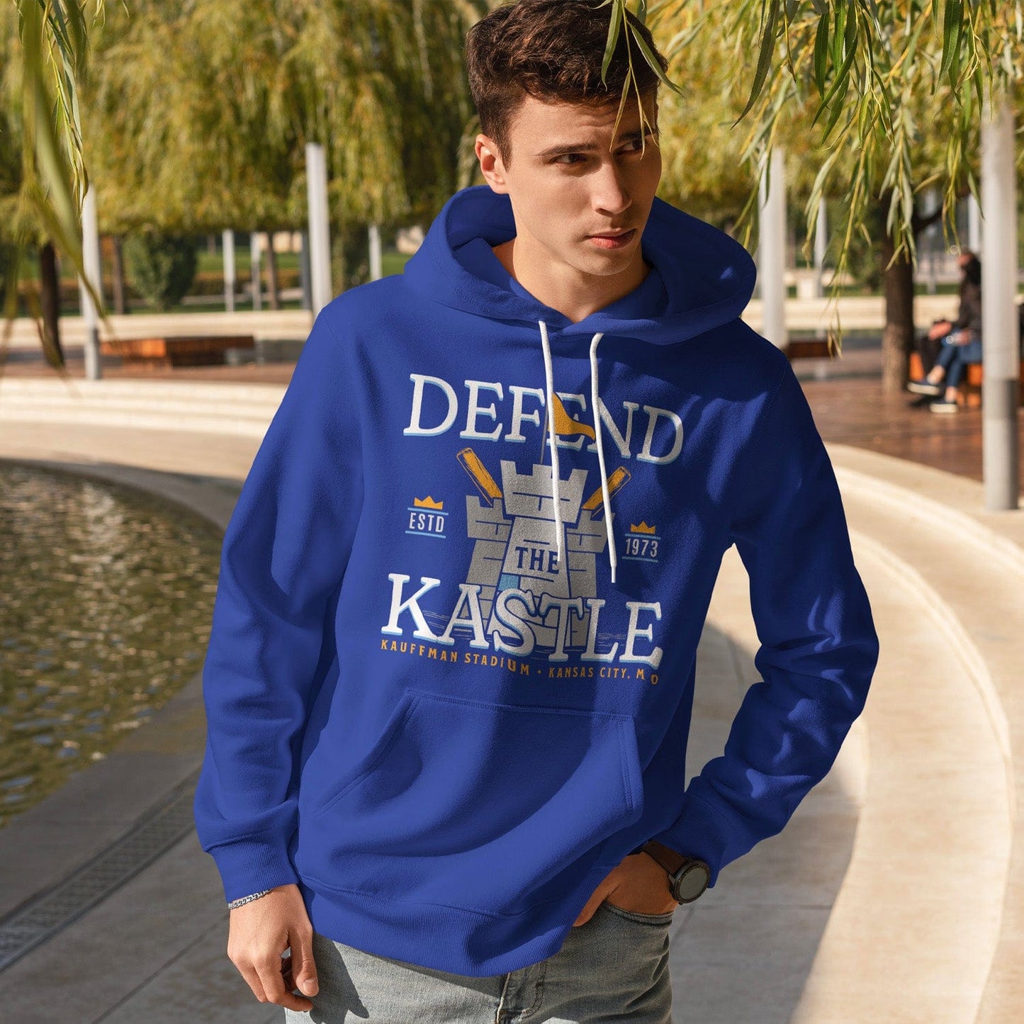 KC Swag Kansas City Royals powder, gold DEFEND THE KASTLE on blue fleece pullover hoodie worn by male model standing near water in public park