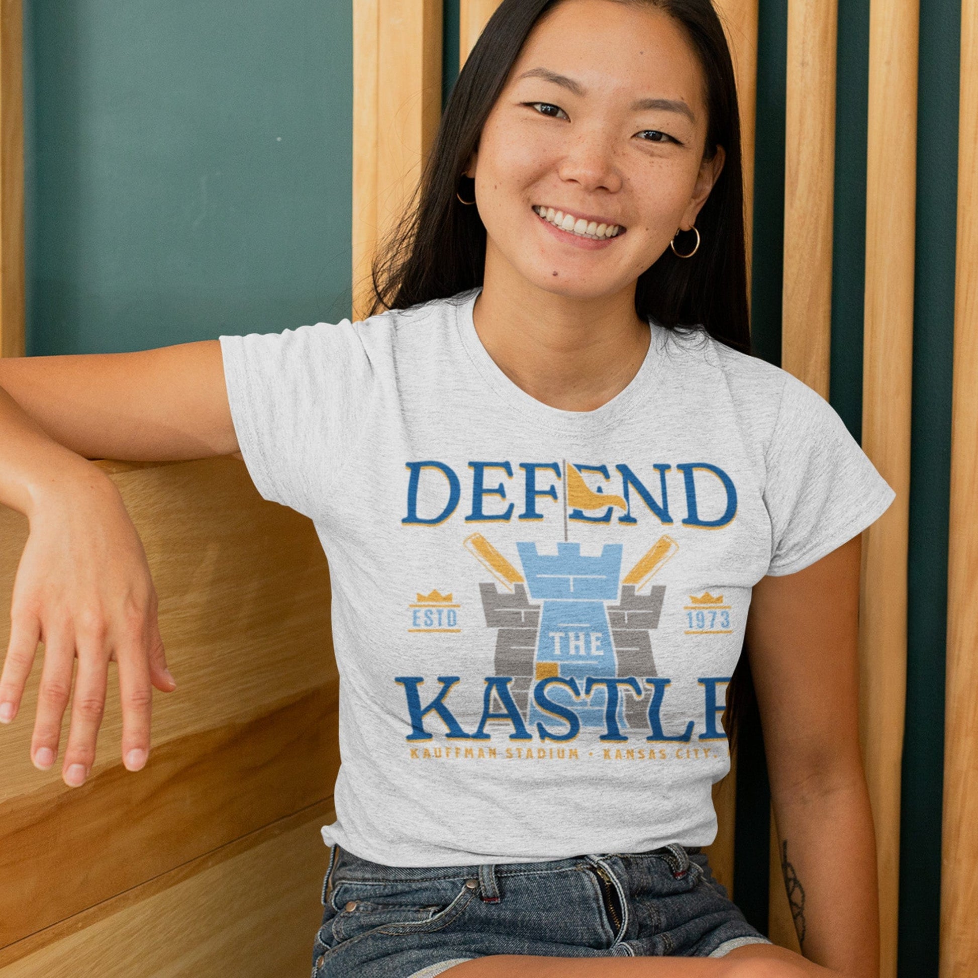 KC Swag Kansas City Royals powder, blue, gold DEFEND THE KASTLE on ash heather grey unisex t-shirt worn by female model sitting in front of wood slat wall