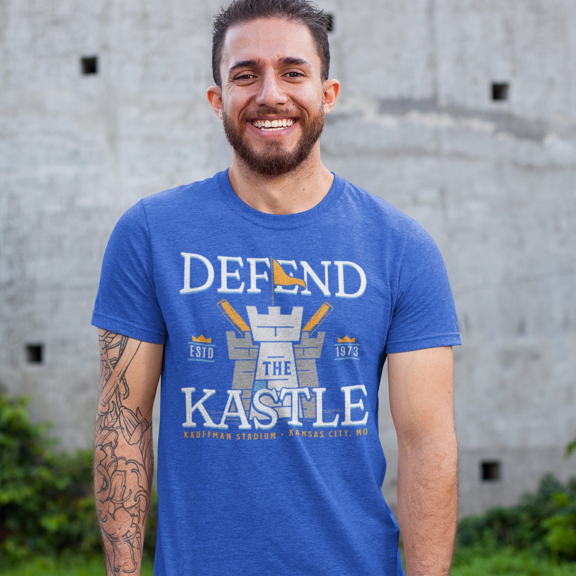 KC Swag Kansas City Royals powder, blue, gold DEFEND THE KASTLE on heather royal blue unisex t-shirt worn by male model standing in front of concrete wall