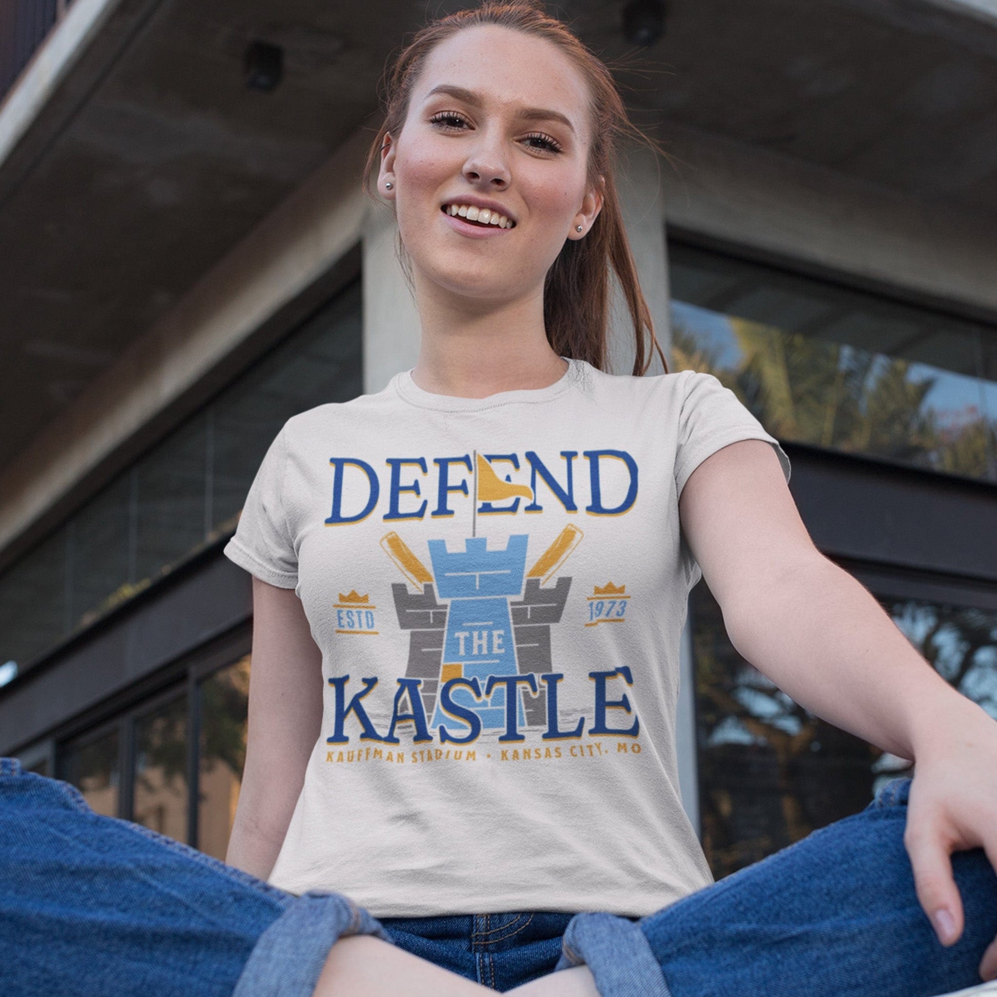 KC Swag Kansas City Royals powder, blue, gold DEFEND THE KASTLE on natural cream unisex t-shirt worn by female model sitting in front of building