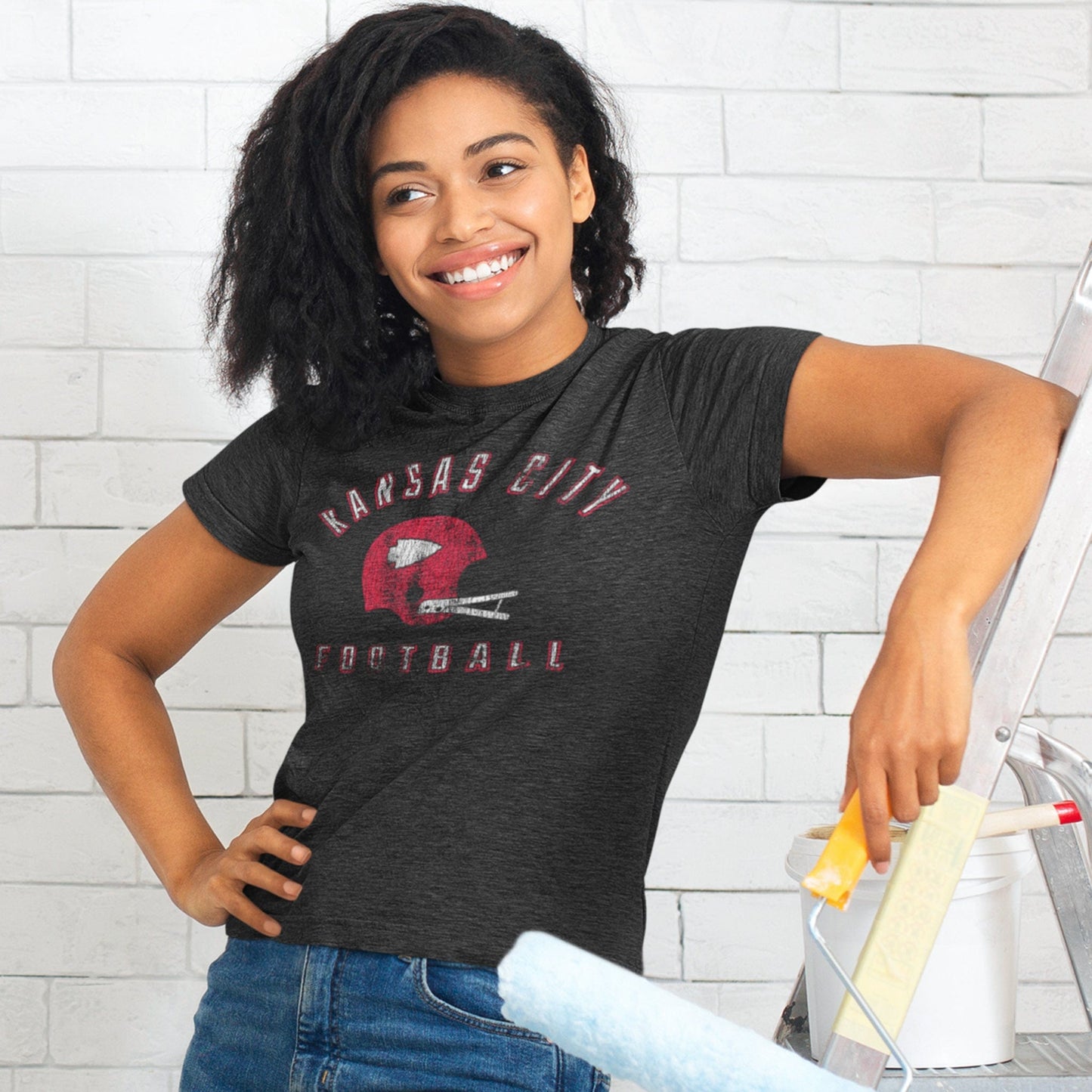 KC Swag Kansas City Chiefs red/white KANSAS CITY FOOTBALL with vintage helmet graphic on dark heather grey t-shirt worn by female model painting white brick wall