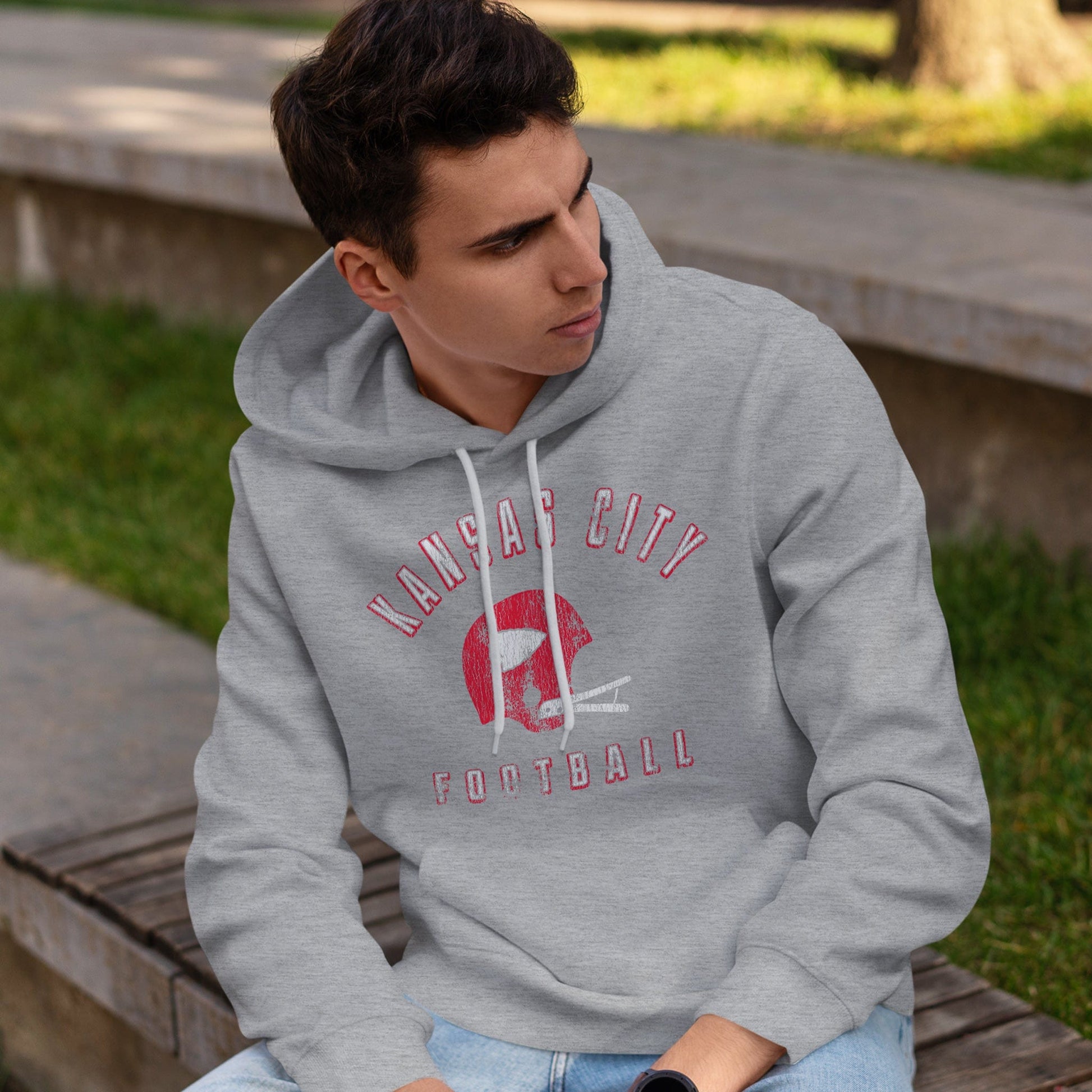 KC Swag Kansas City Chiefs red/white KANSAS CITY FOOTBALL with vintage helmet graphic on athletic heather grey fleece pull-over hoodie worn by male model sitting on bench in park