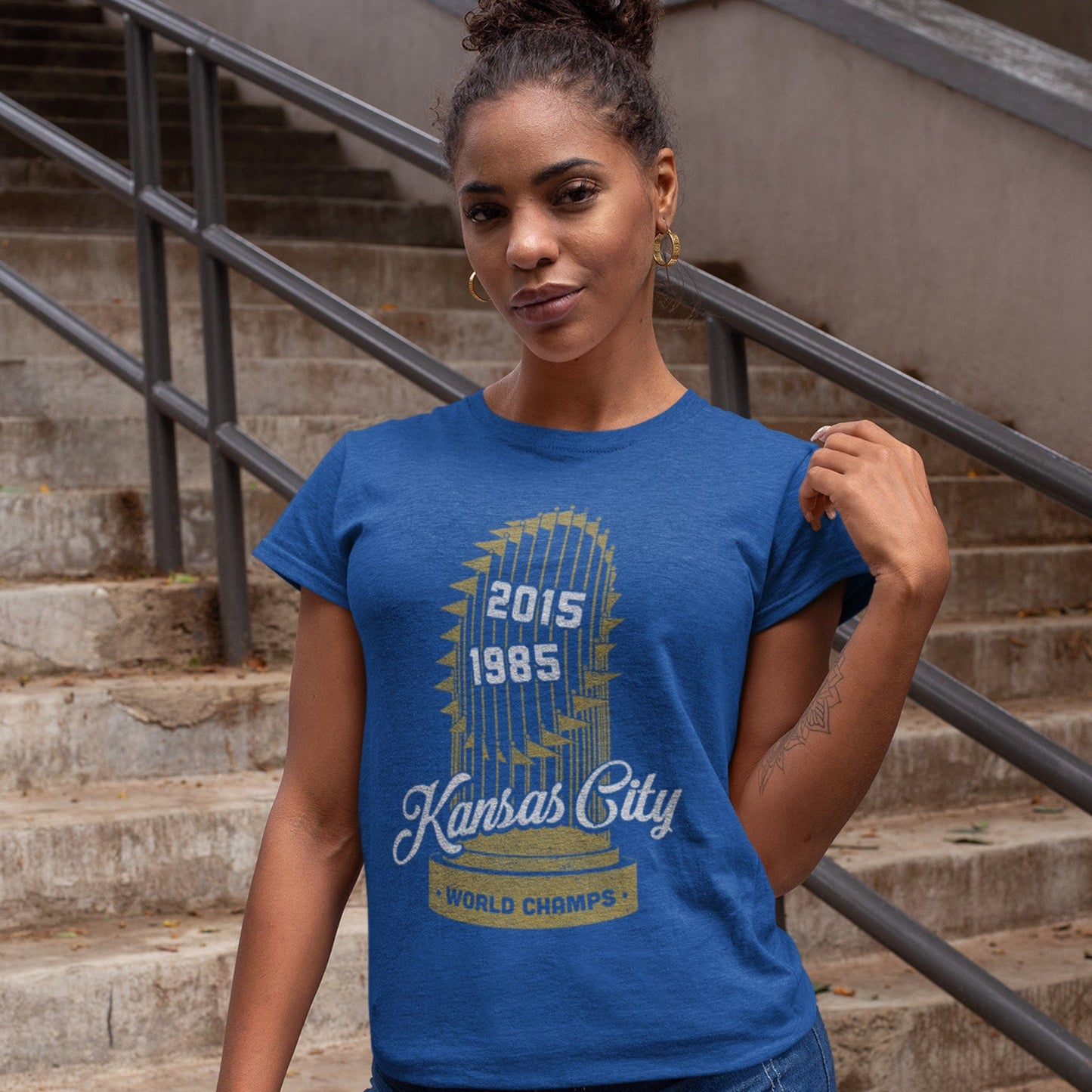 KC Swag Kansas City Royals white KANSAS CITY 2015 1985 with gold WORLD SERIES TROPHY on heather royal blue t-shirt worn by female model standing on concrete staircase